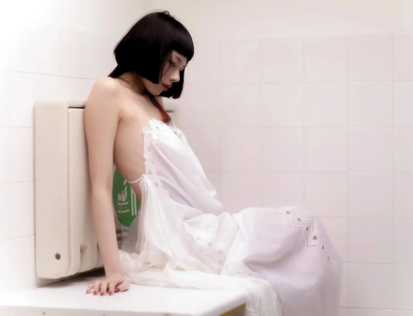 A woman sitting on a bathroom counter, partially covered with a white sheet, with a wall-mounted sign indicating a restroom, in a minimalist, white-tiled bathroom.