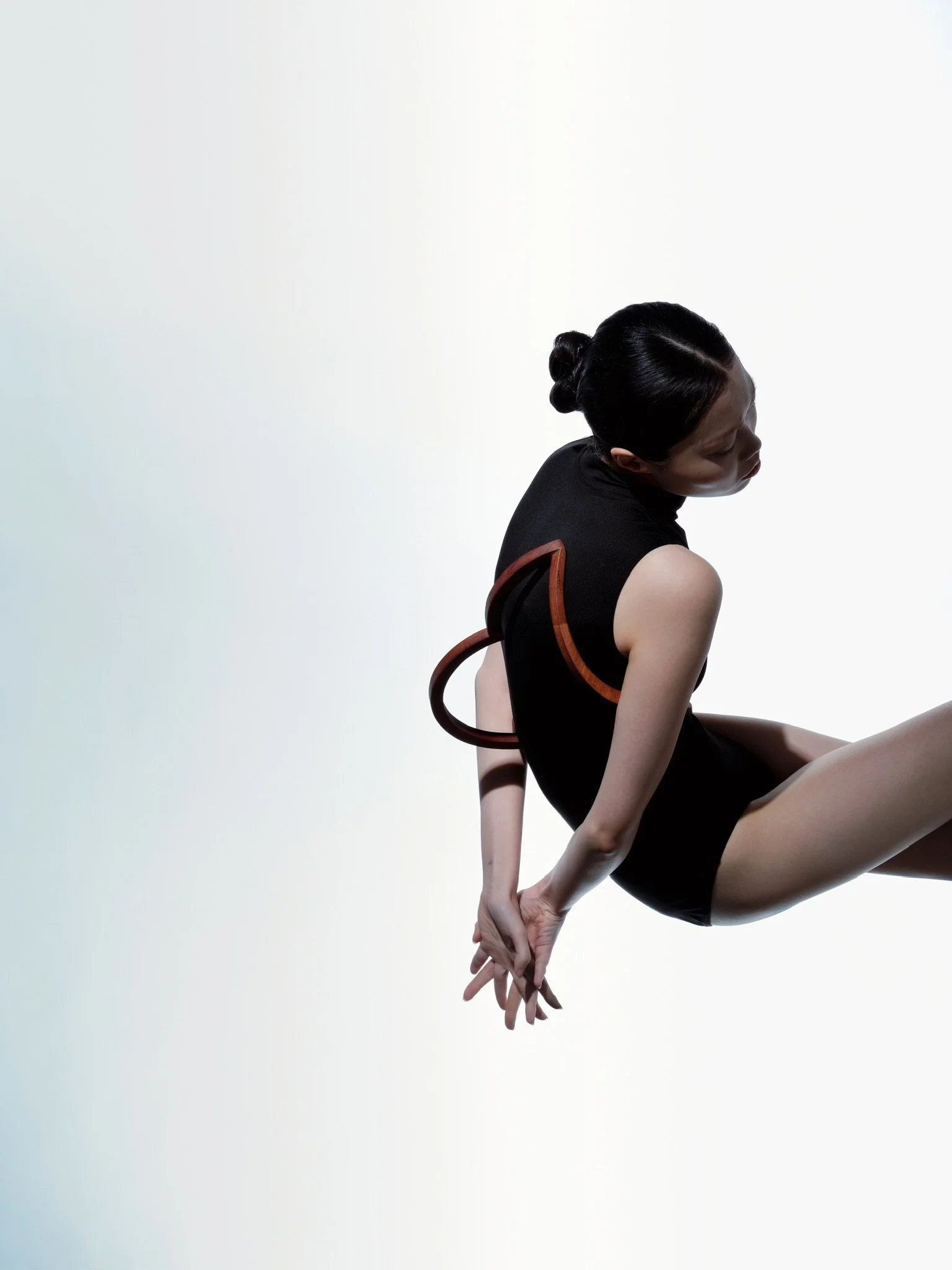 A woman with black hair styled in a bun, wearing a black sleeveless top, seated against a plain light background, with her head tilted downward and her hands joined behind her back.