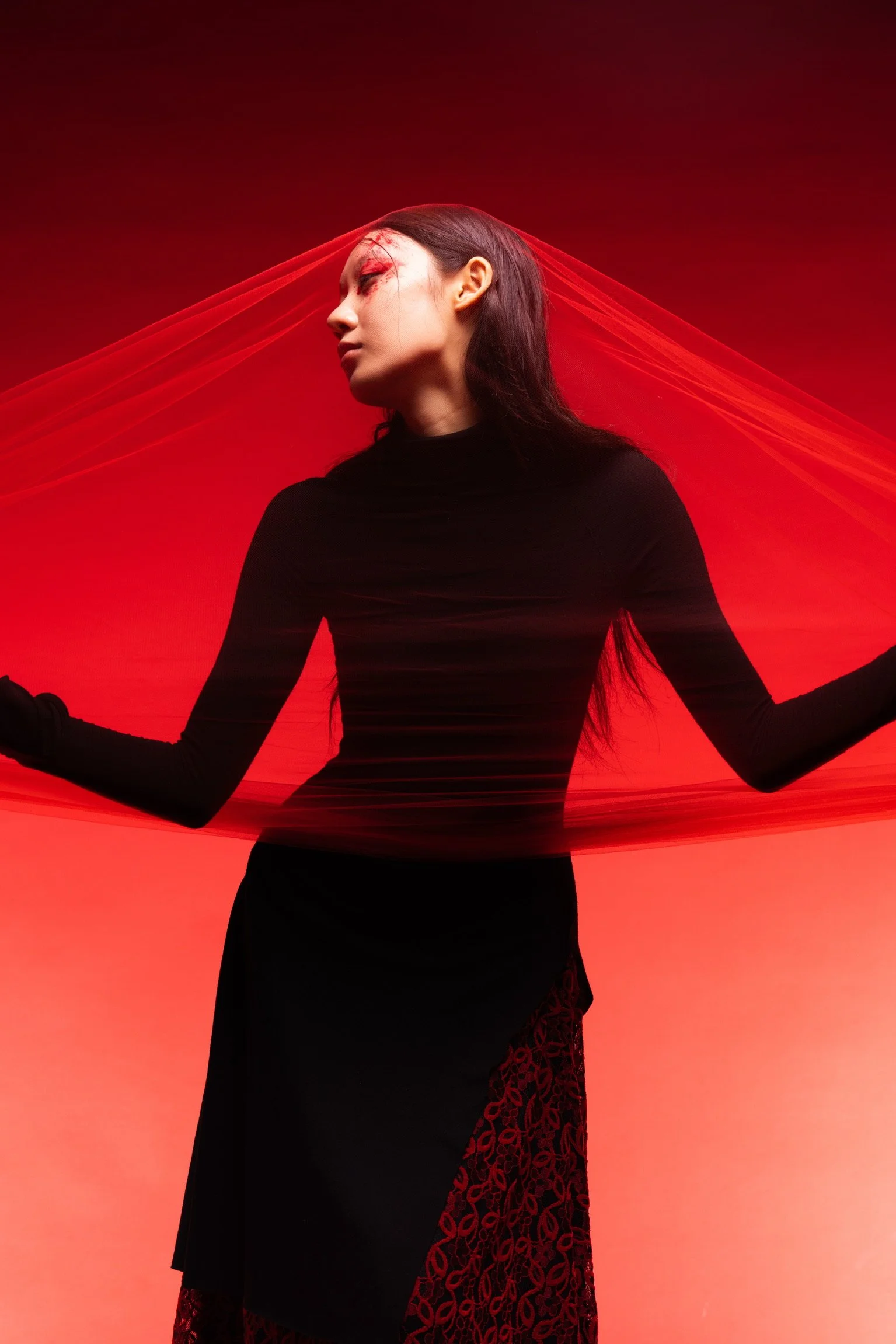 A woman in a black dress with red lace details stands against a red background, with a red fabric draped around her head and arms in a dramatic pose.