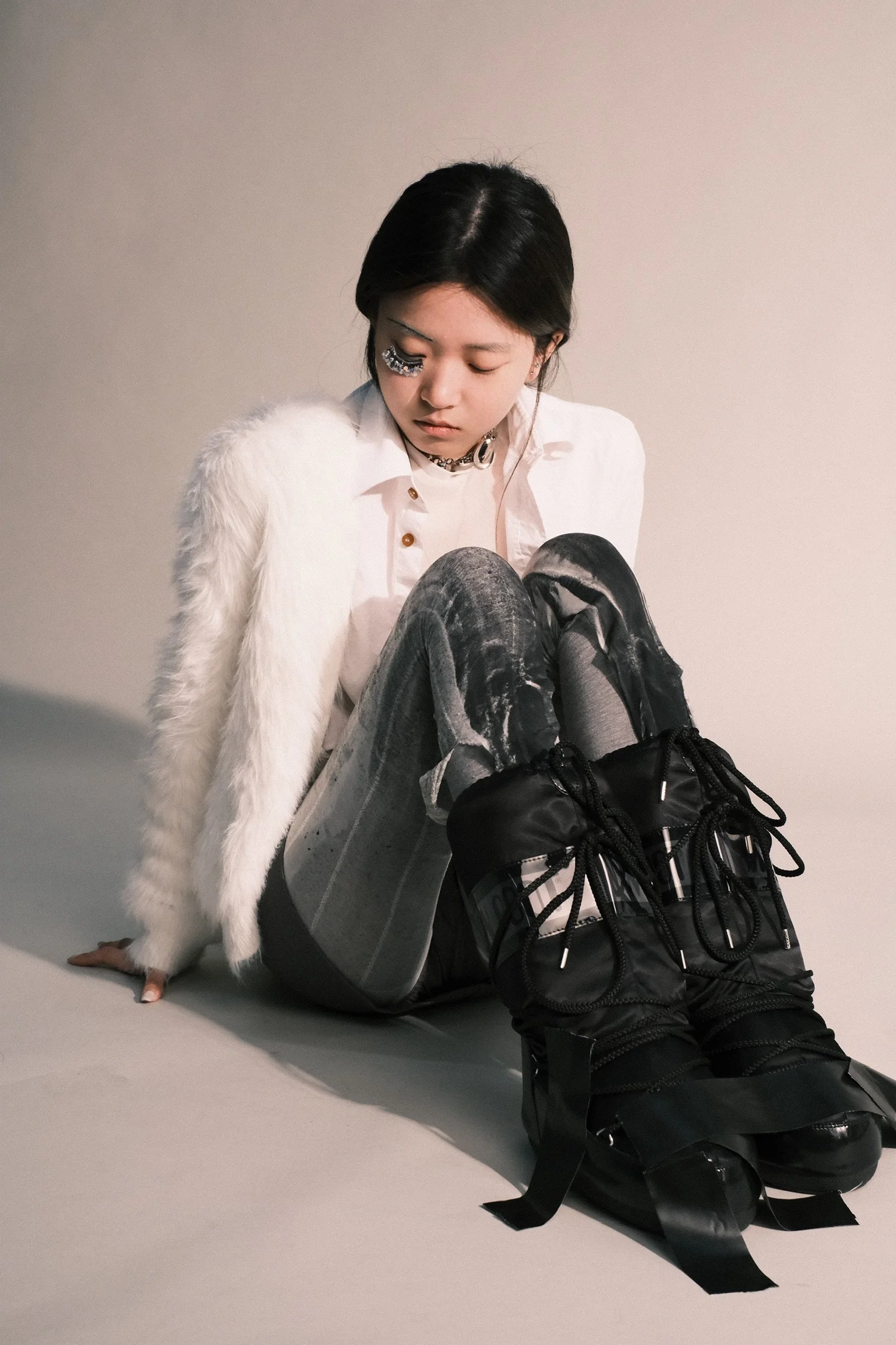 A woman with black hair and a white furry coat is sitting on the floor with her back against a wall. She is dressed in a white shirt, ripped jeans, and black sneakers, with a backpack on the ground in front of her. Her head is tilted downward and she has one eye closed with decorative face gems around it.