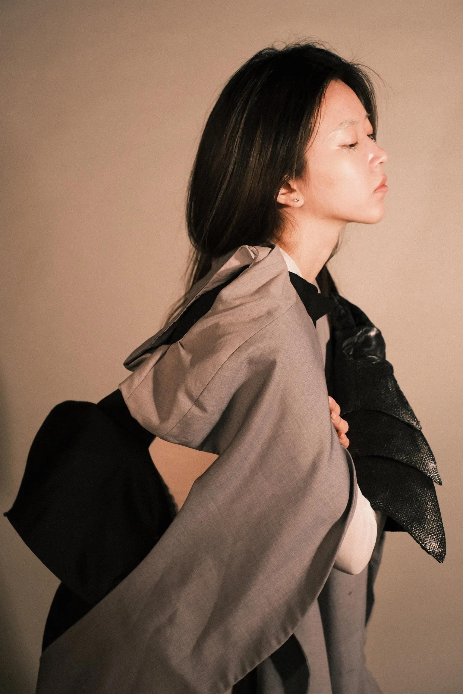 A woman with dark hair and light makeup, dressed in layered clothing, is shown in profile against a plain background.
