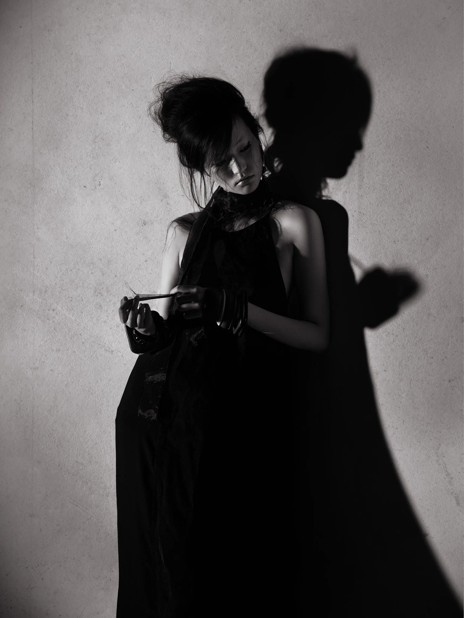 Black and white photo of a woman with styled hair wearing dark clothing, holding an object in her hand, standing against a textured wall. Her shadow is cast on the wall behind her.