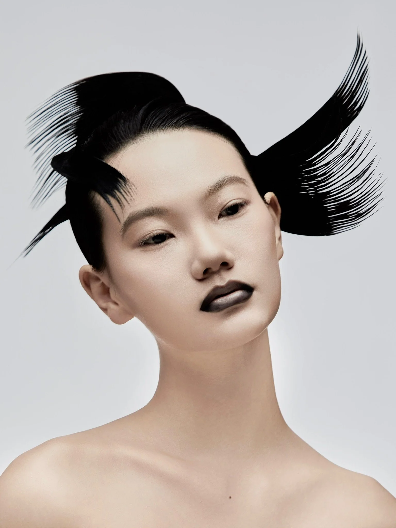 A woman with a high-fashion hairstyle incorporating large, black, feathered hair extensions, minimal makeup including black lipstick, and a bare shoulder against a plain light gray background.