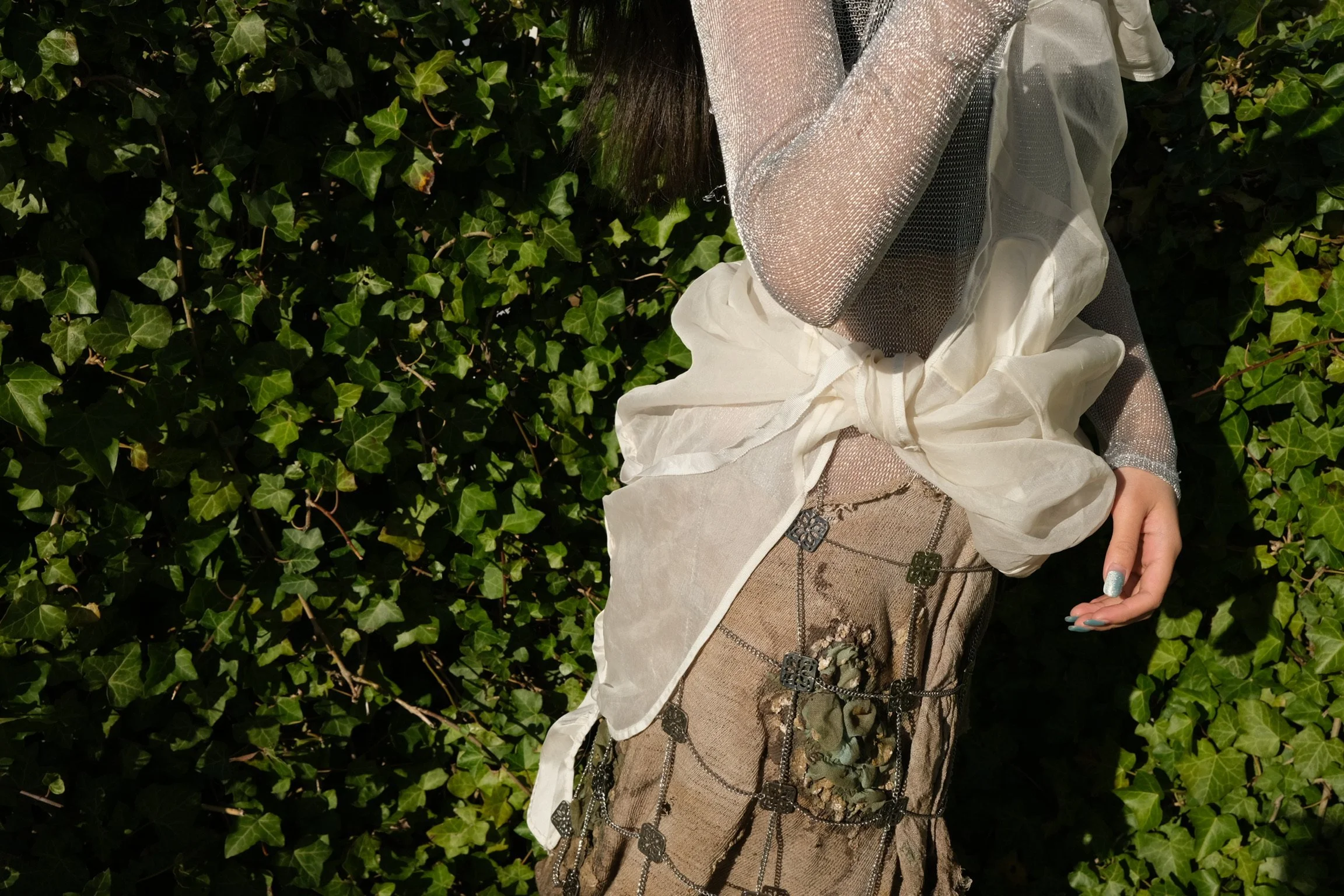 Close-up of a person wearing a shiny silver jacket and layered clothing, standing against a background of green ivy leaves, with one hand showing silver and blue nails.