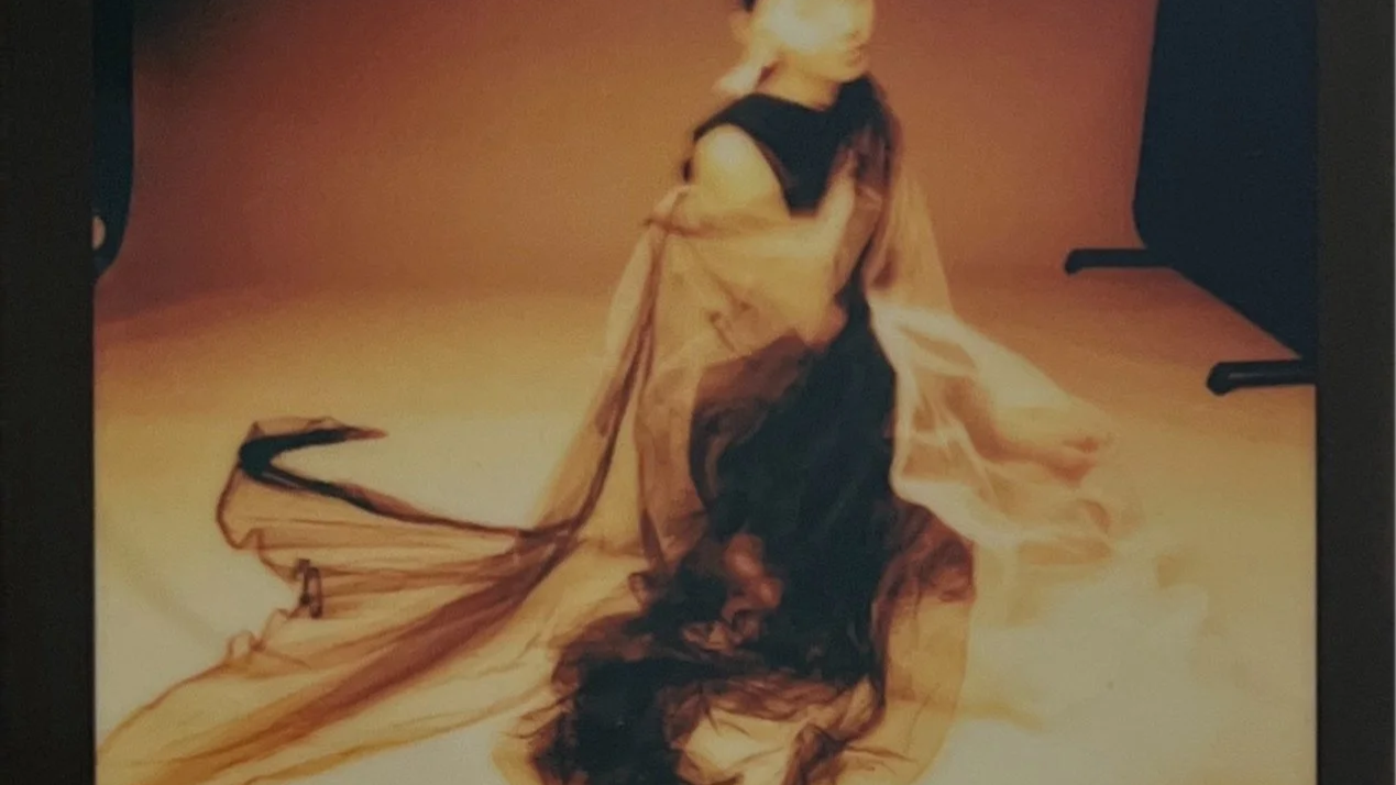 A person with long blonde hair in a black dress, draped in a sheer, flowing tan-colored fabric, seated on a tan-colored floor with a brownish background.