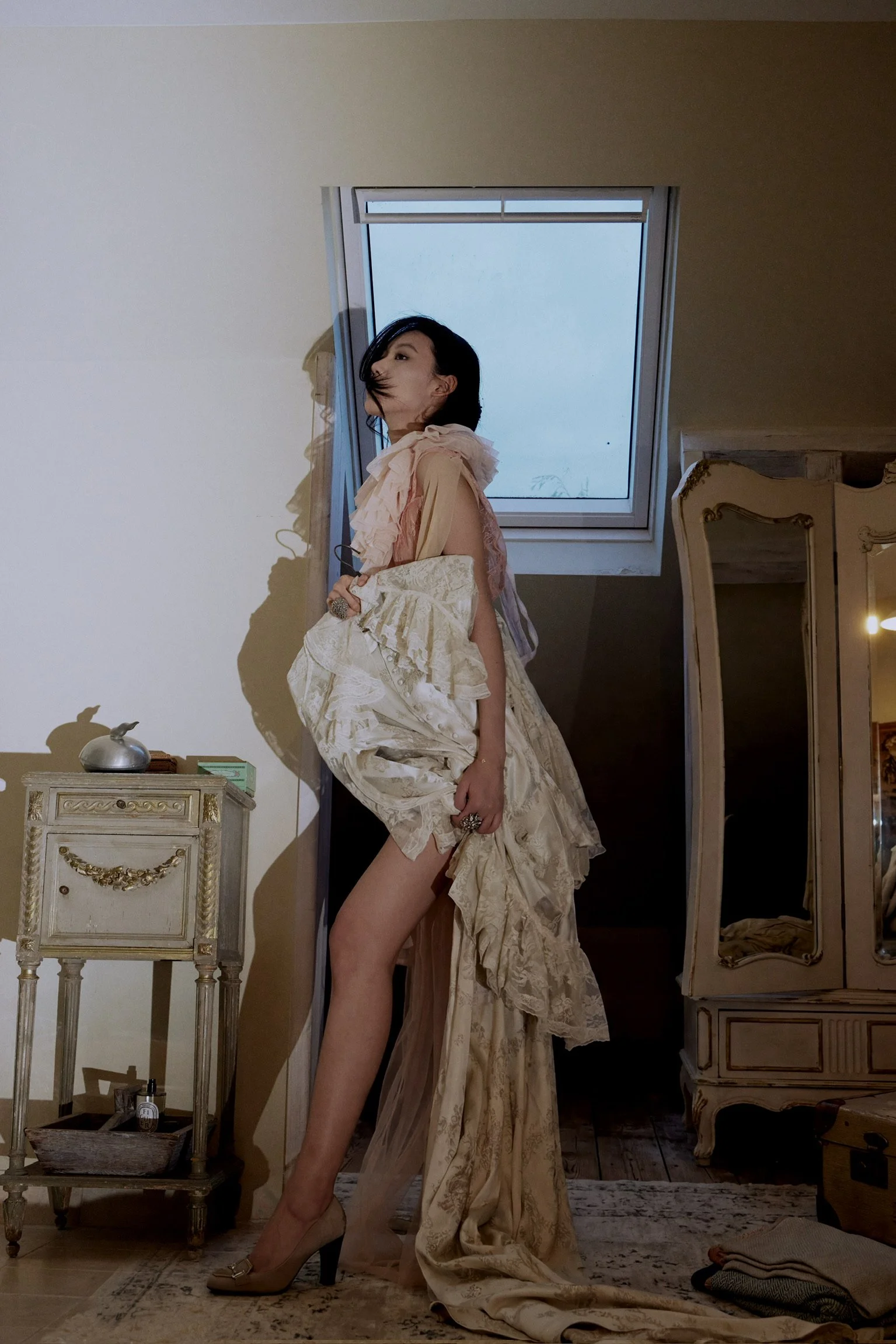 A woman with black hair stands in a room, partially covered by an ornate, cream-colored dress that drapes over her shoulders and cascades down. She is holding a part of the dress with her left hand, exposing her left leg. She wears high heels and has accessories on her fingers. The room has vintage furniture, a window in the ceiling with snow outside, and a mirror reflecting parts of the room.