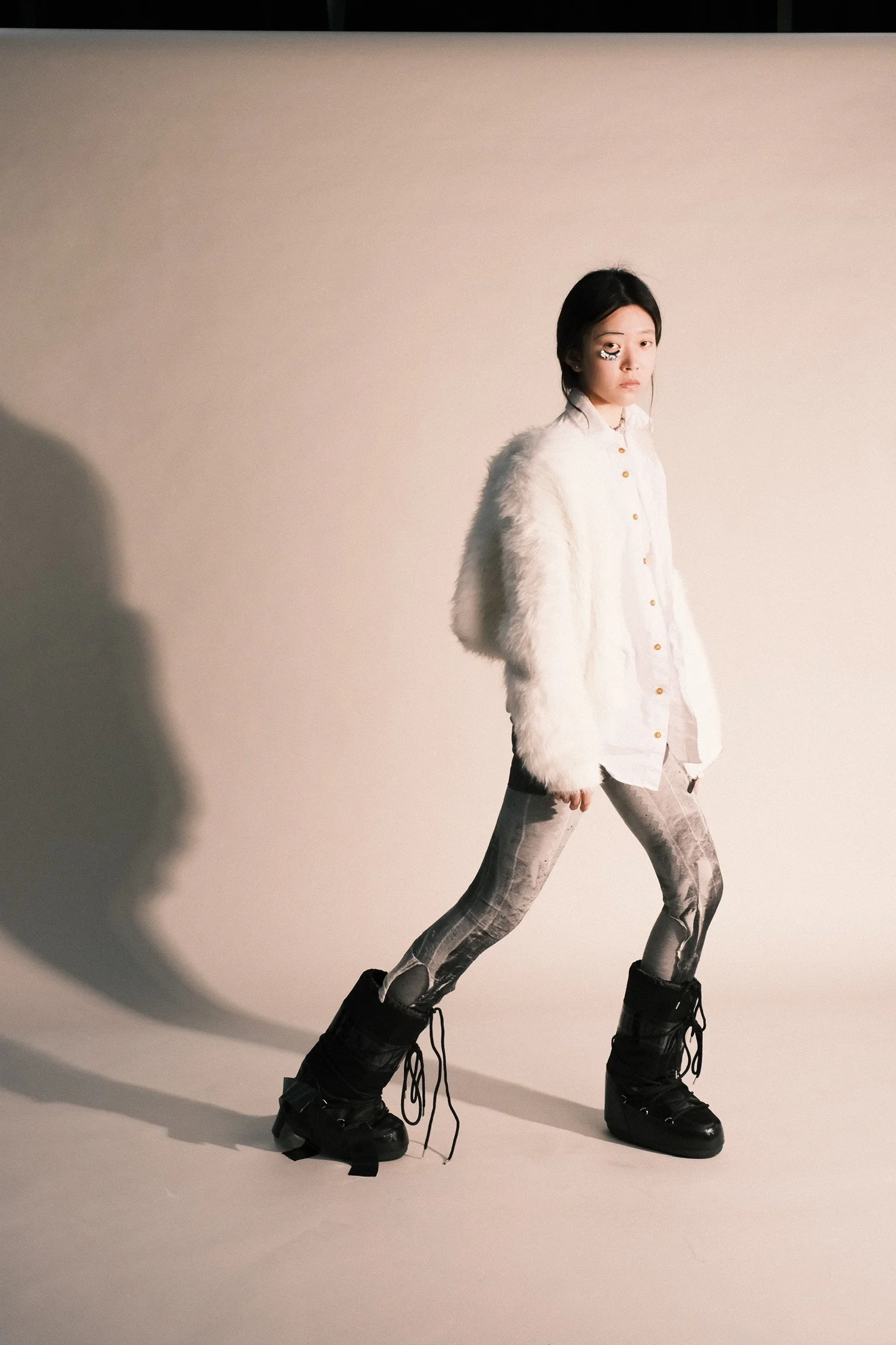 A young woman with short black hair and an eye patch on her right eye, dressed in a white shirt, a fluffy white jacket, patterned leggings, and black combat boots, walking with a dynamic pose against a plain background.