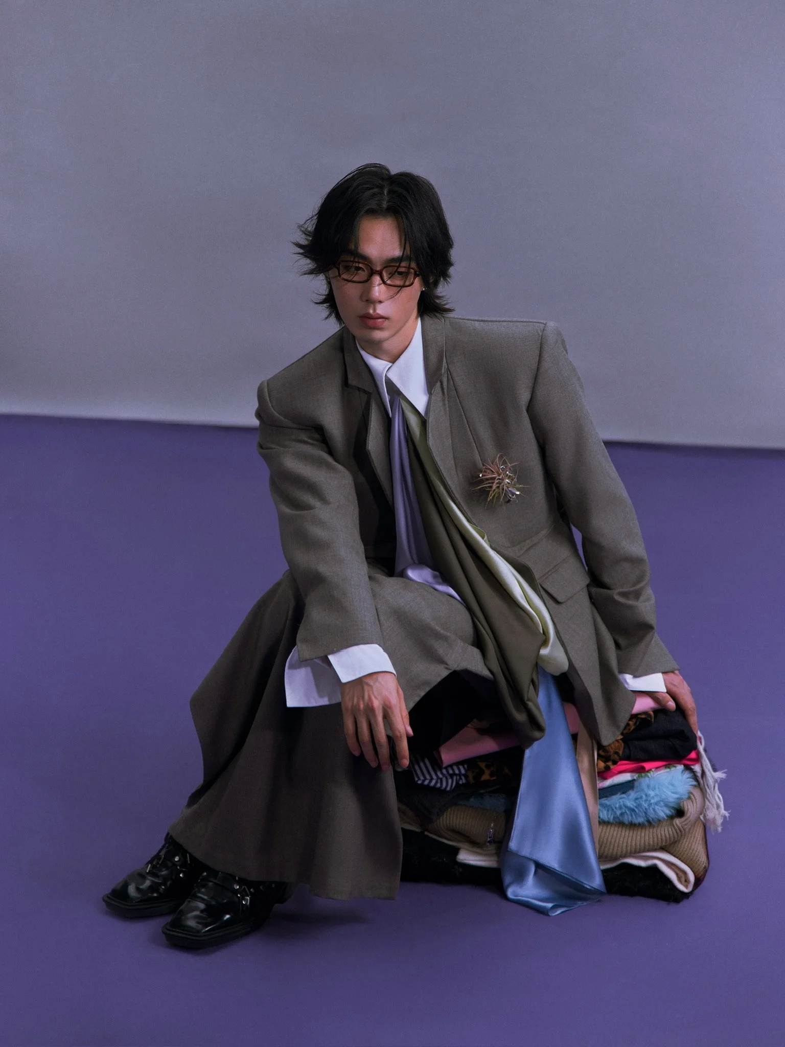 A young man with black hair, glasses, and wearing a gray suit over a white shirt and purple tie. He is sitting on the floor with a pile of colorful clothes and bags next to him, against a purple background.