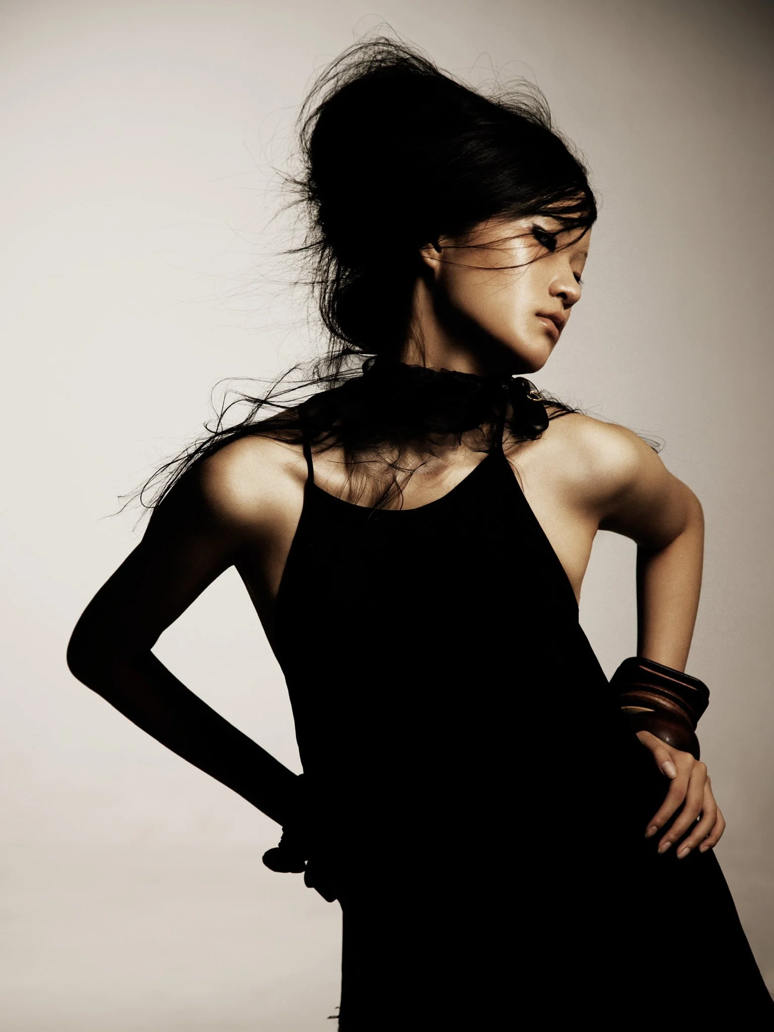 A woman with dark hair and a black dress poses against a plain background, with her head turned to the side and one arm bent.