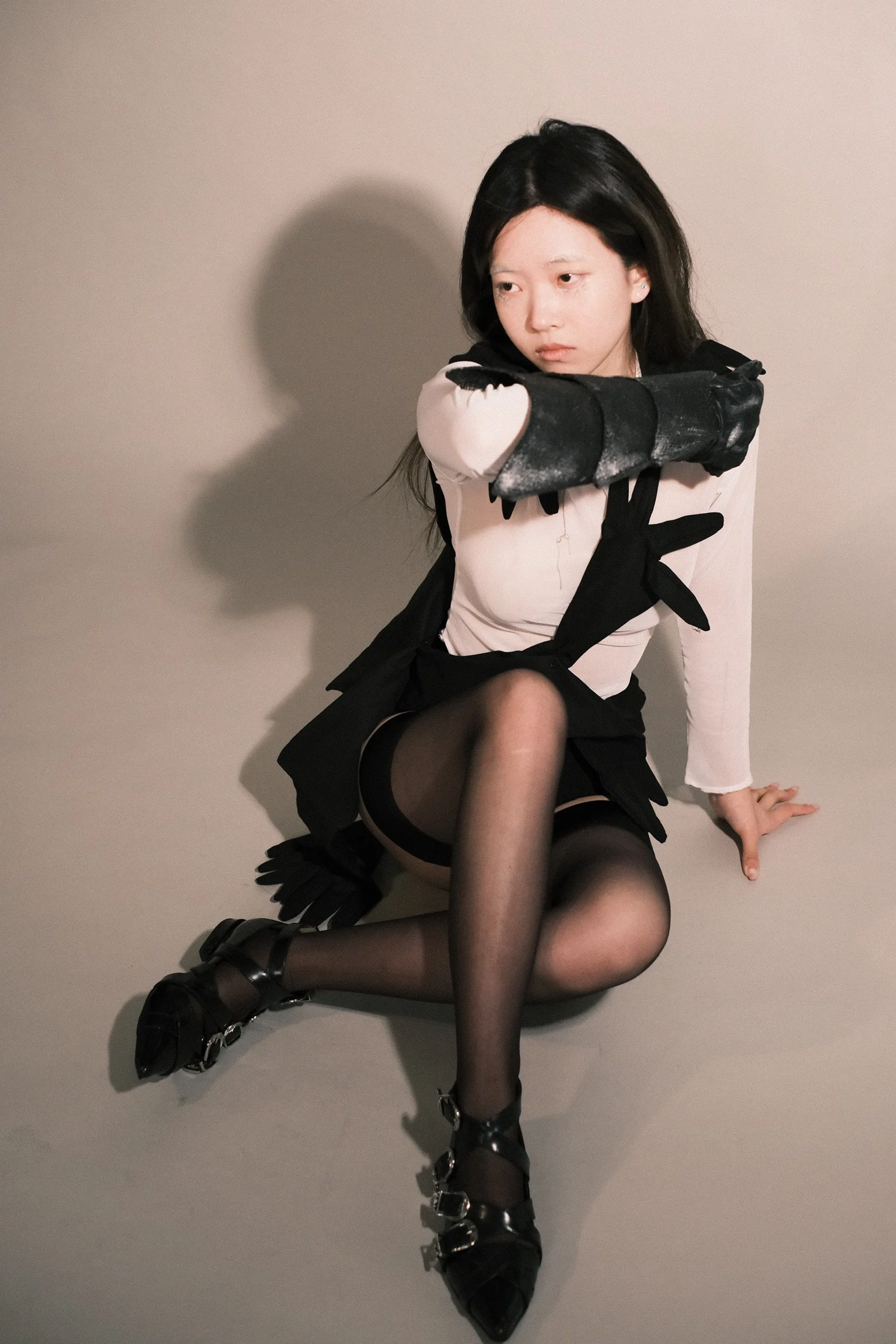 A young woman with black hair sitting on the floor against a plain wall. She is wearing black sheer stockings, black heeled shoes with chains, a black and white outfit, and long black gloves. She is posing with her legs crossed, arm resting on her knee, and looking pensively to the side.
