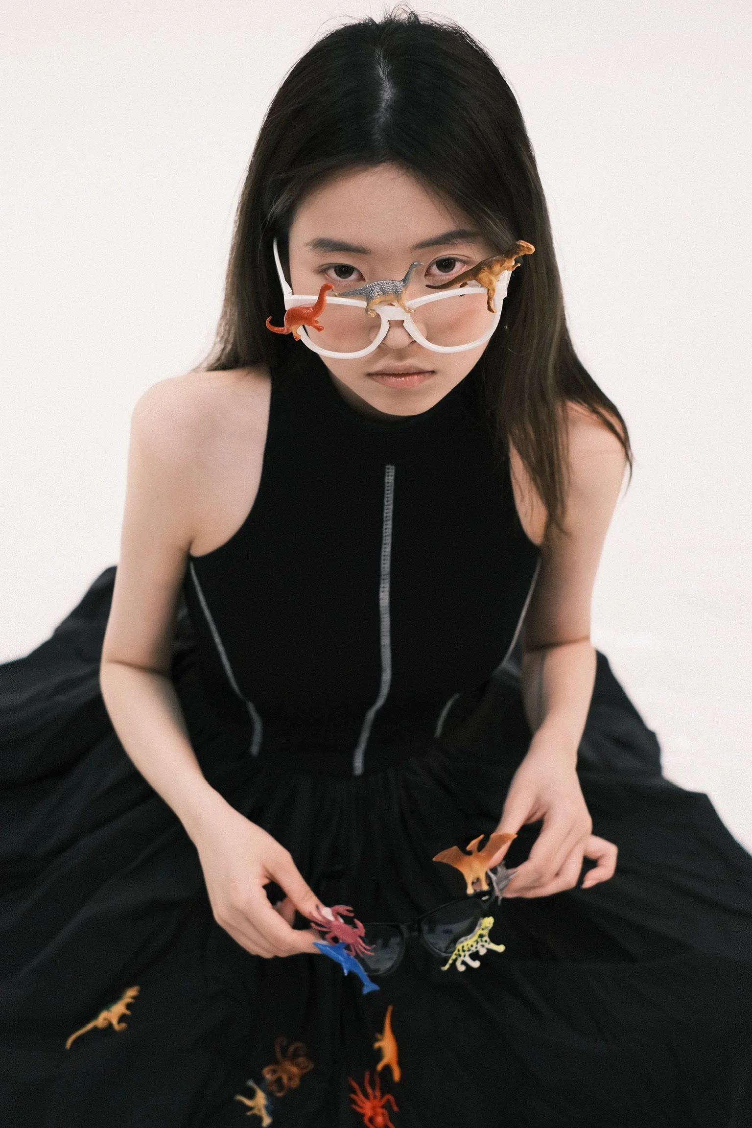 A young woman with long dark hair wearing glasses and a sleeveless black top, sits against a plain white background. She is playing with small, colorful plastic dinosaur toys spread on her lap and a black surface.