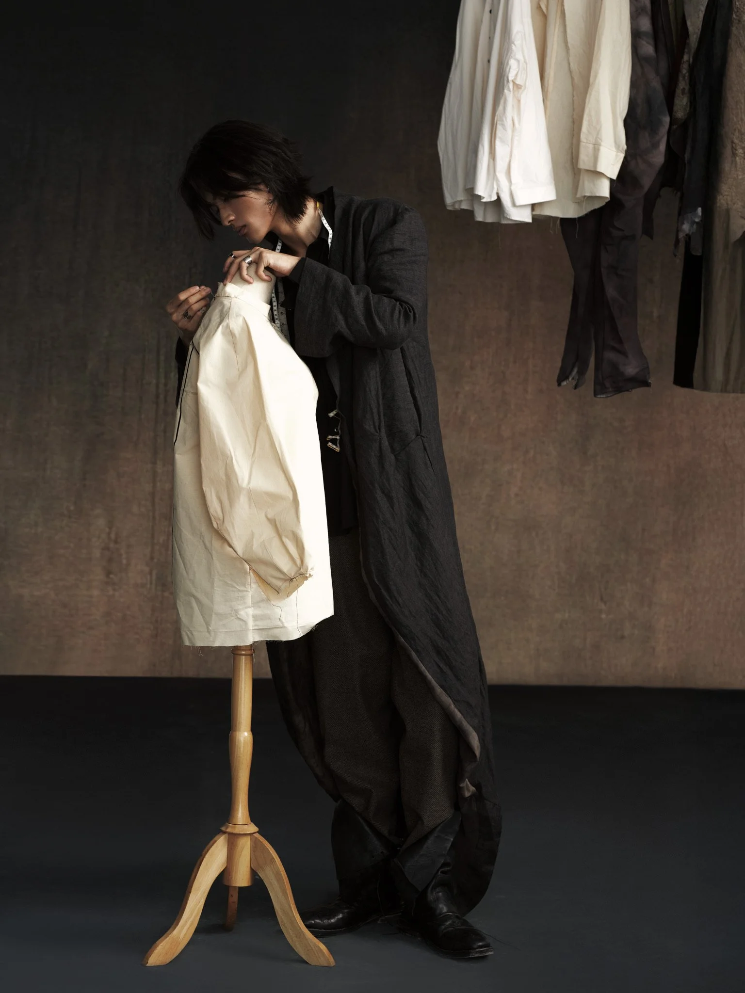 A person in dark clothing working on a white garment on a mannequin, with various clothes hanging in the background.
