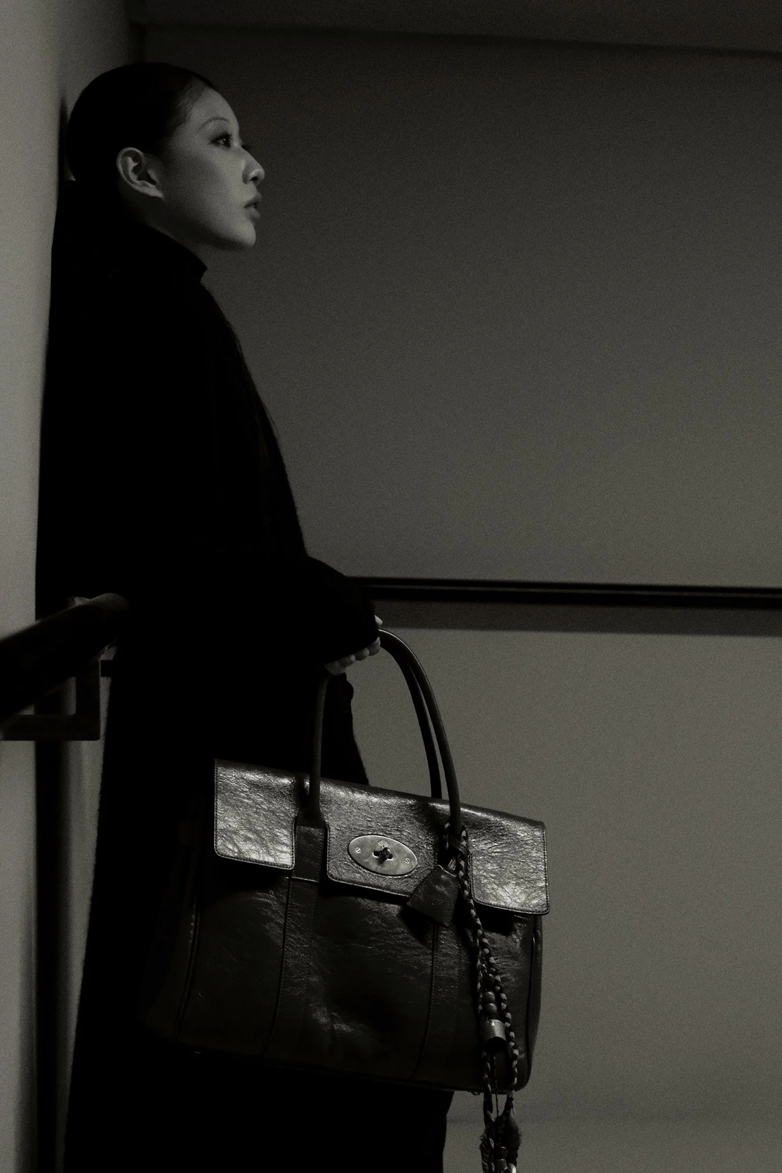 A woman in profile with dark hair tied back, standing against a wall in a dimly lit room, holding a large handbag with a chain strap.