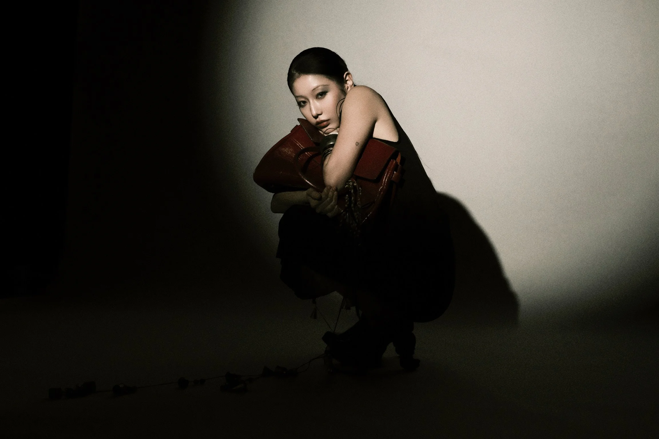 A woman with short dark hair and tattoos on her arms, sitting with her head resting on her arms, holding a large brown purse, in a dark environment with a plain light-colored background.