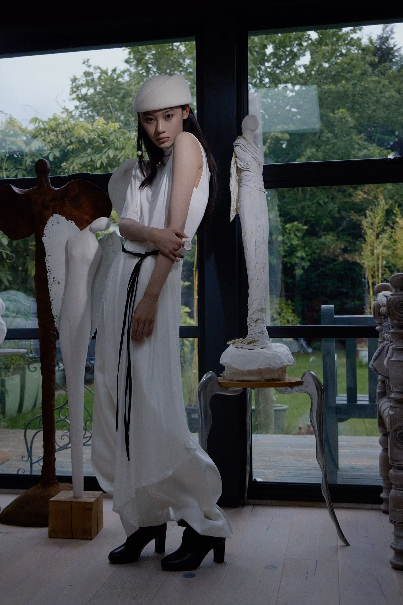 A young woman with long dark hair, wearing a white dress, white beret, and black heels, standing inside a room with large windows showing green trees outside. There are various sculptures and art pieces around her, including a tall white abstract sculpture on a stool, a wooden sculpture of a cow, and a column with fabric wrapped around it.