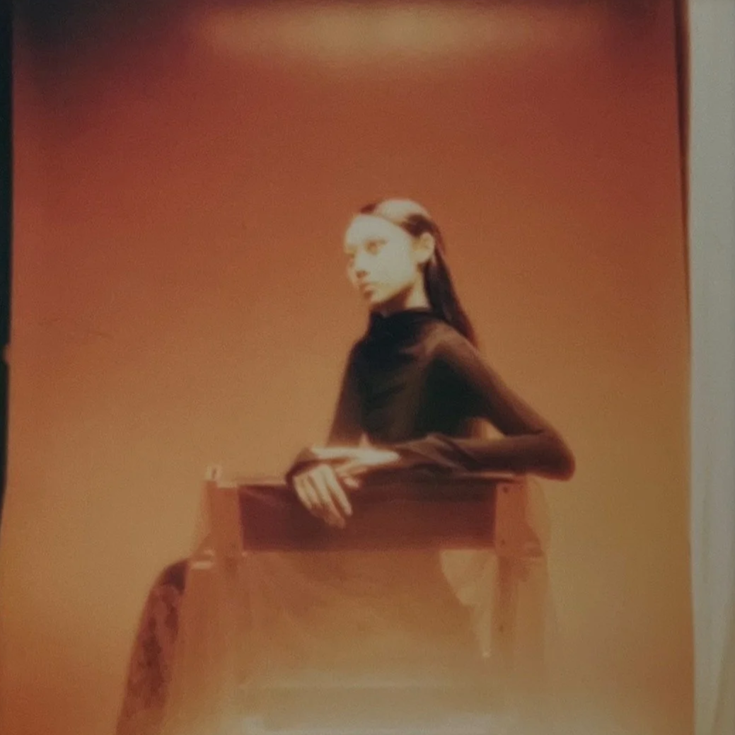 A woman with long dark hair, wearing a black top, leans on a wooden surface in a dimly lit room with reddish ambient lighting.