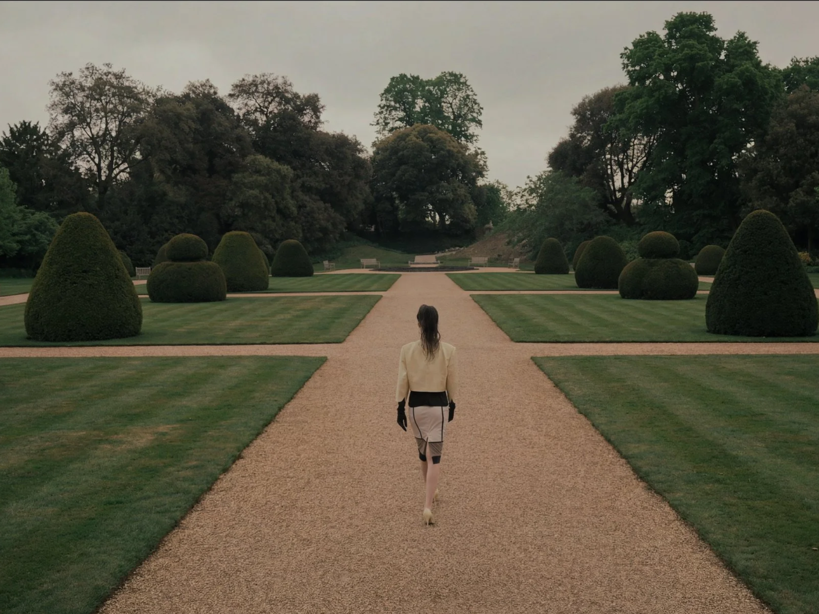 A woman dressed in a white jacket and beige skirt walking on a gravel path in a formal garden with manicured lawns and shaped bushes, with trees in the background.