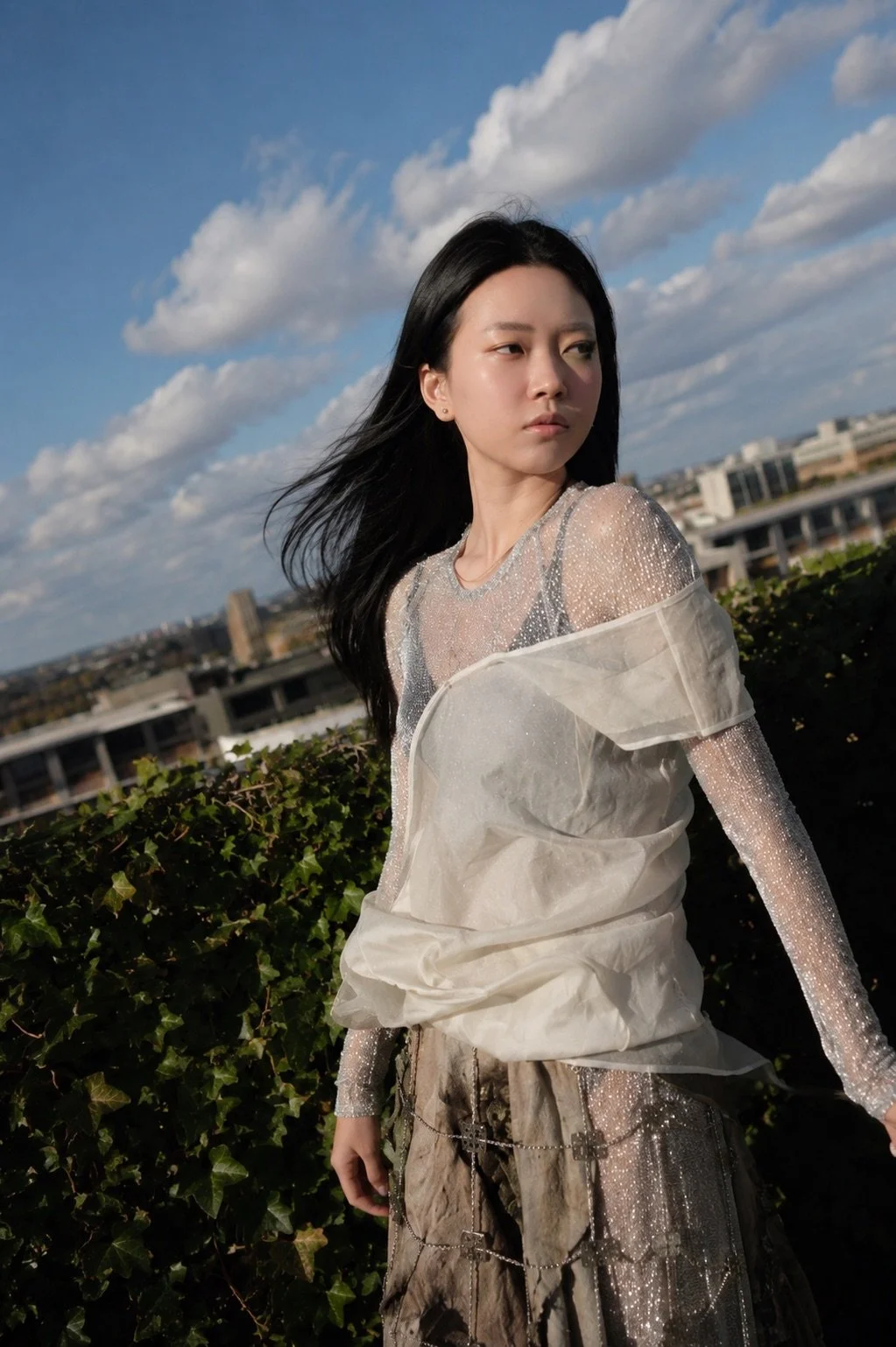 Young woman with long dark hair standing outdoors on a rooftop or terrace, facing left, with a cityscape and partly cloudy sky in the background.