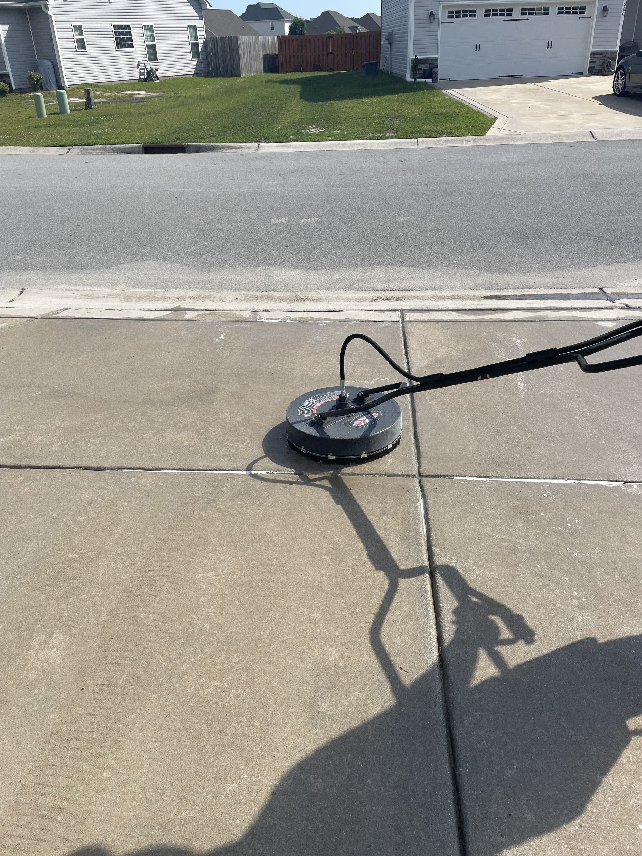 A concrete driveway with a pressure washing machine in the foreground, casting a shadow on the surface. The background includes a street, a grassy lawn, and houses.