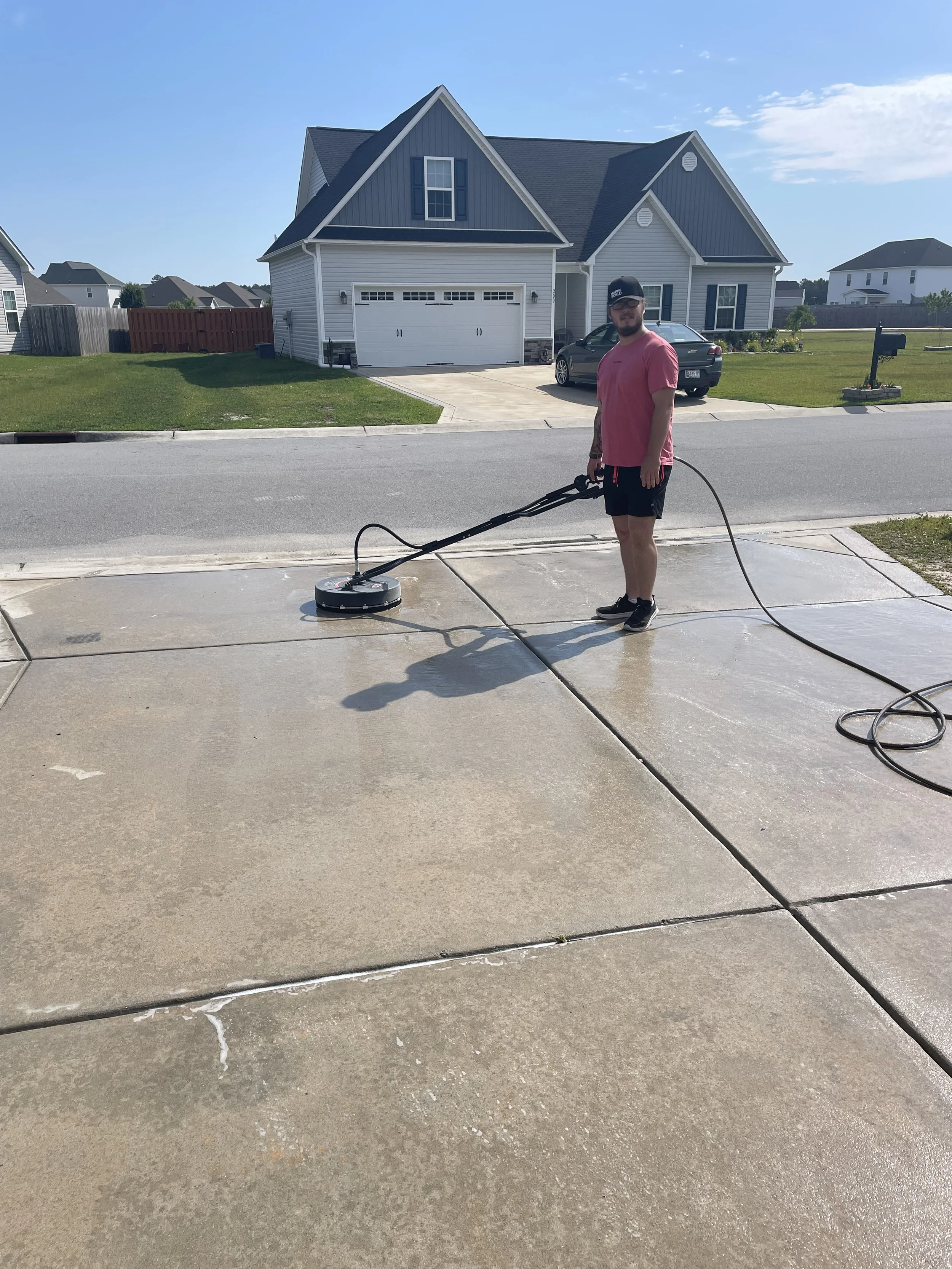 A man using a robotic floor cleaner on a driveway in front of a house on a sunny day.