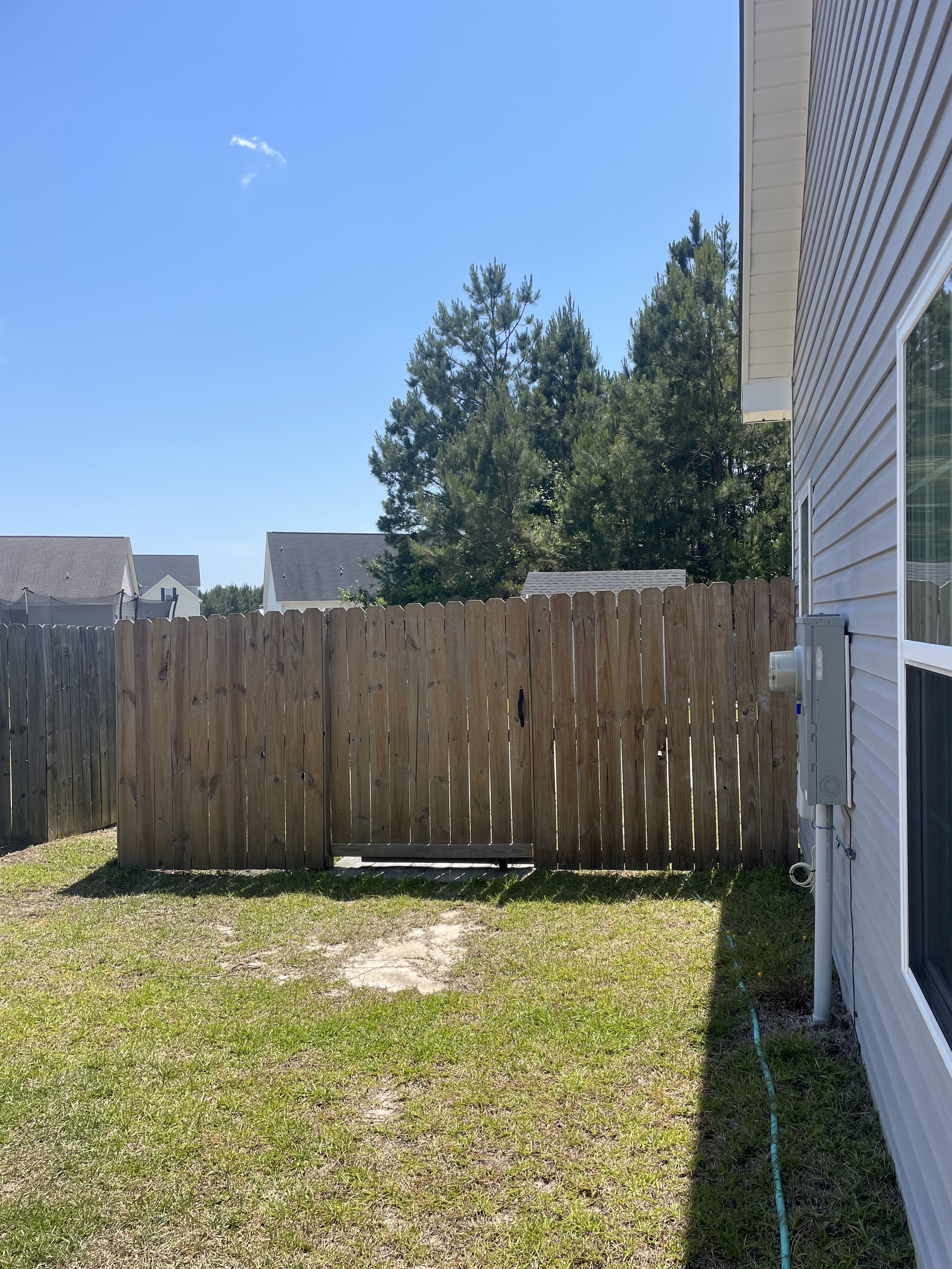 Backyard with grassy lawn, wooden fence, white house siding, electrical boxes, large trees, neighboring houses, and a blue sky with a small cloud.