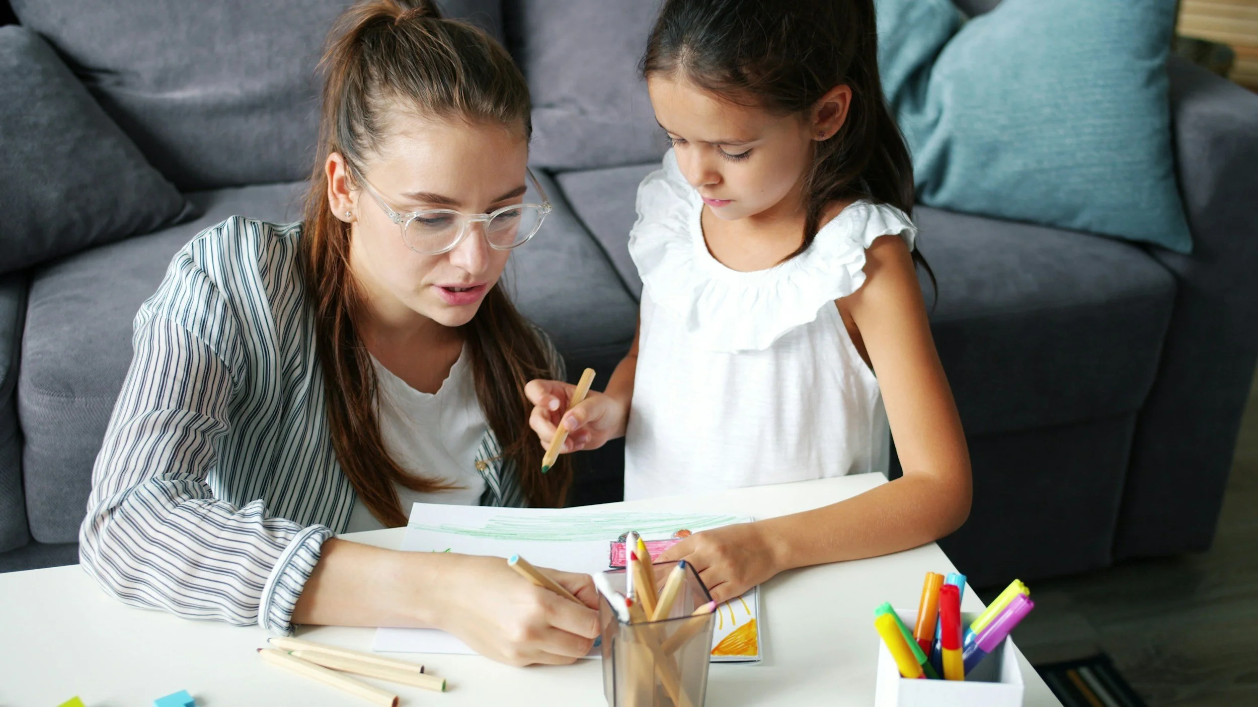 A young woman and a young girl sit at a table, working together on a drawing or coloring project. The woman wears glasses and a striped shirt, while the girl wears a white top and has dark hair. Various colored markers and pencils are on the table, and a container of markers is also visible.