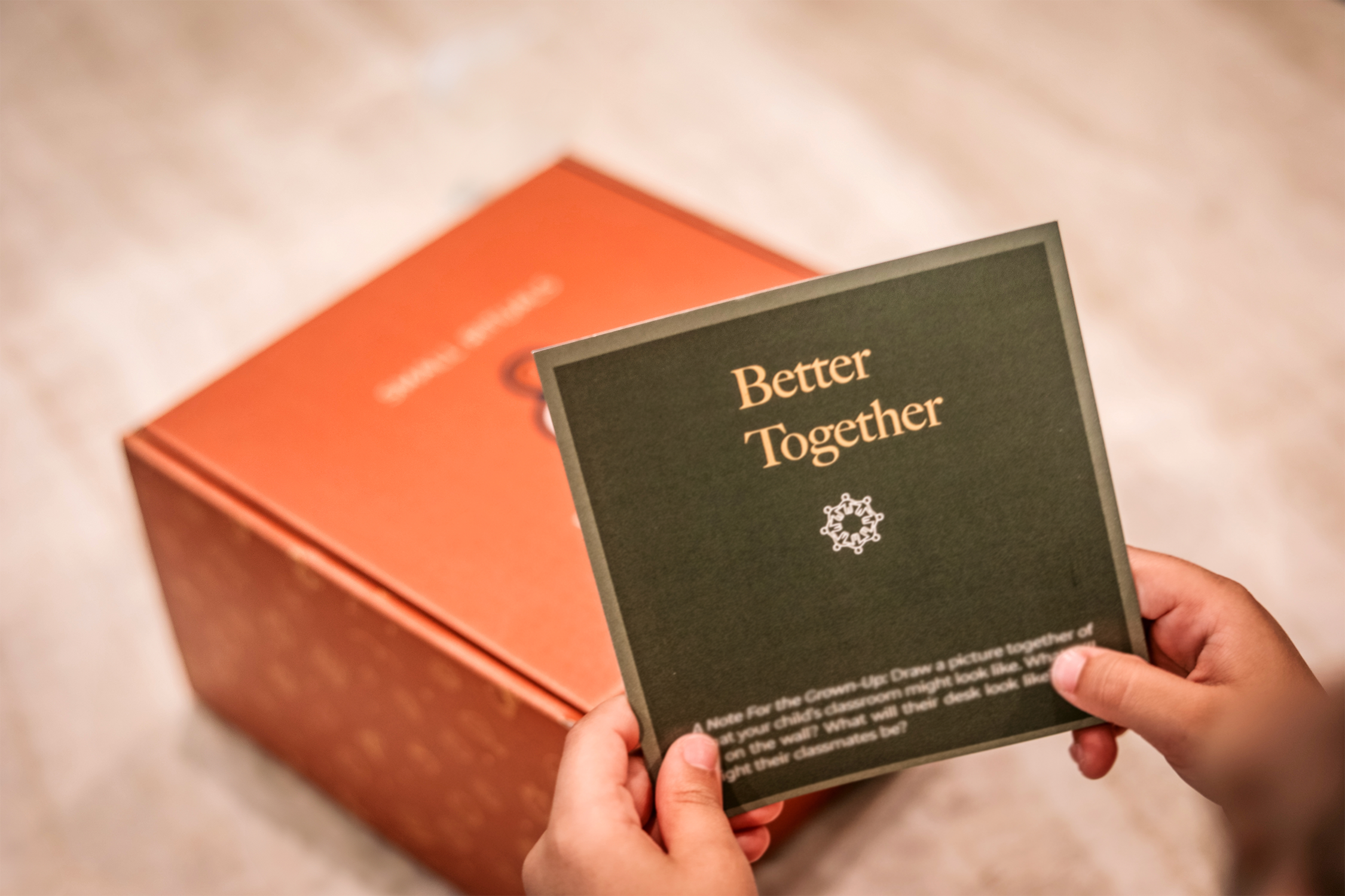 Person holding a black card displaying the text 'Better Together' with a white symbol below, near a red box on a beige surface.