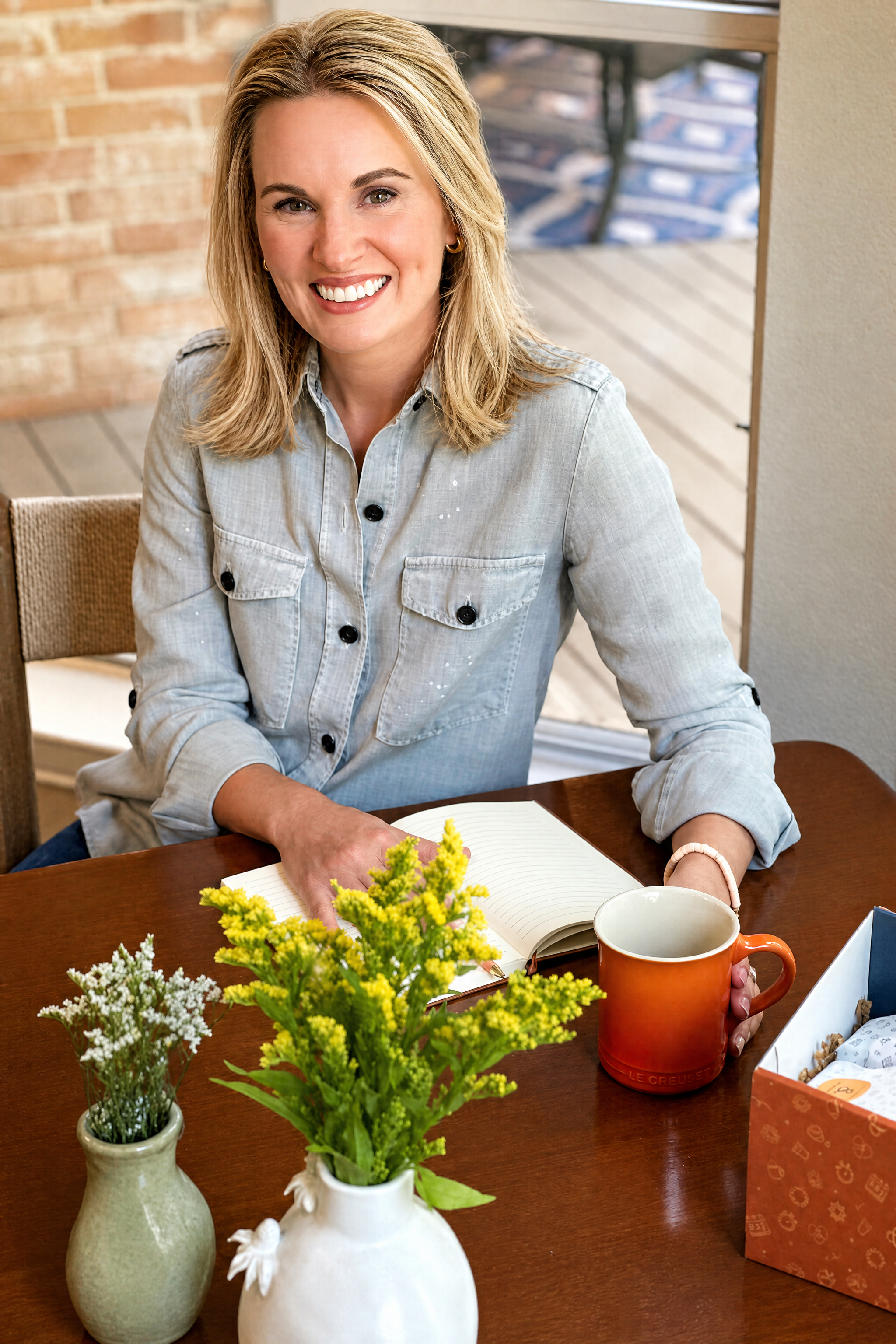 Jessica Vidrik, founder and CEO of Milk & Coffee Kids sitting at a wooden table, smiling, with a notebook, orange mug, and flowers in vases in front of her, in a cozy indoor setting.