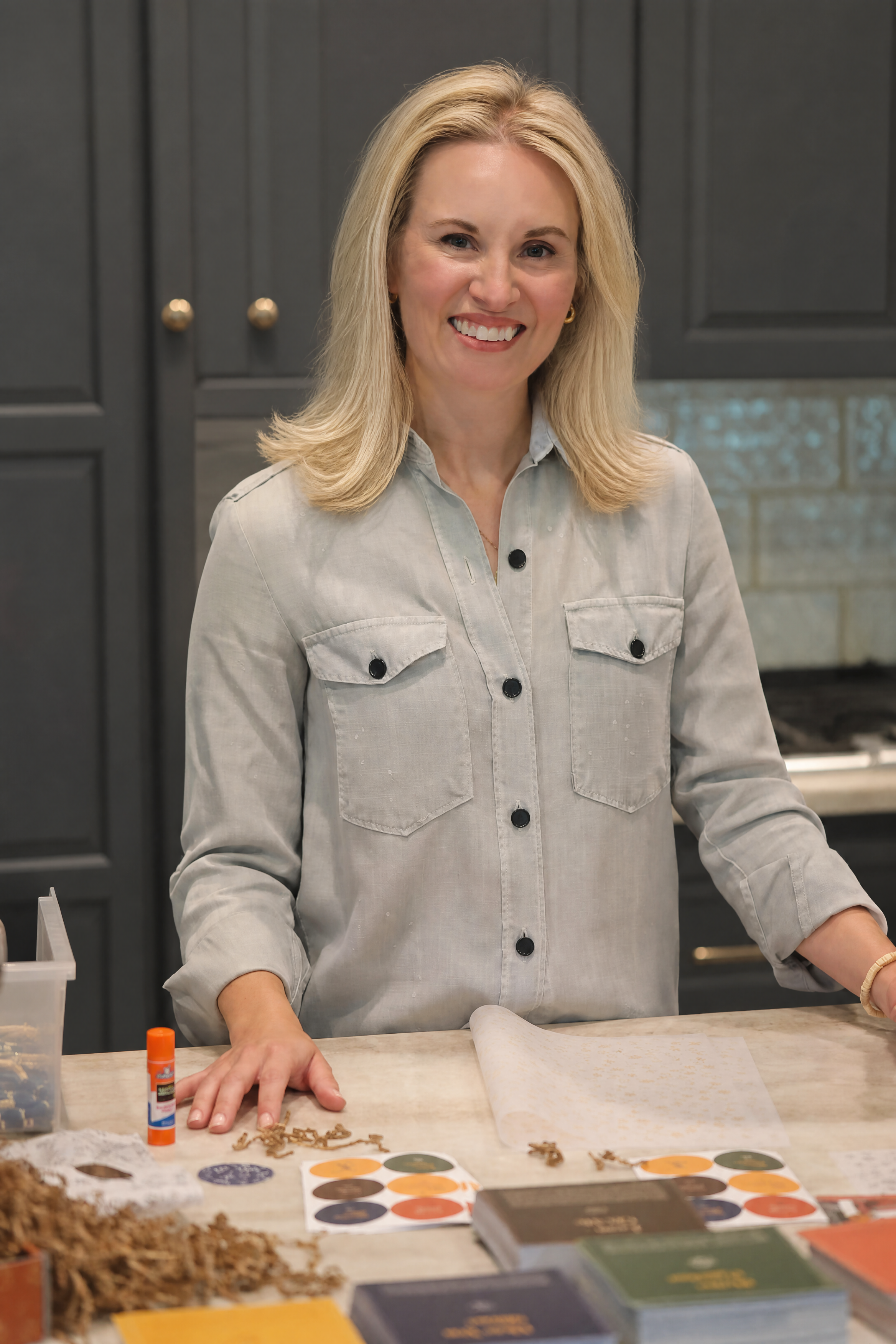 Jessica Vidrik in a light gray shirt sits at her home workspace with Milk & Coffee Kids kits' supplies, including glue, paper, and color palettes, in a kitchen with dark gray cabinets and beautiful backsplash.