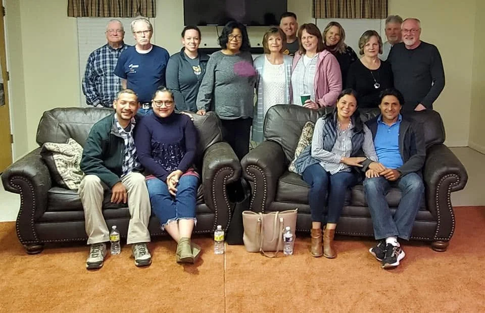 A group of fifteen people posing together in a living room, some sitting on two dark leather sofas and others standing behind them. There is a beige carpet, a cozy curtain, and a TV on the wall in the background.