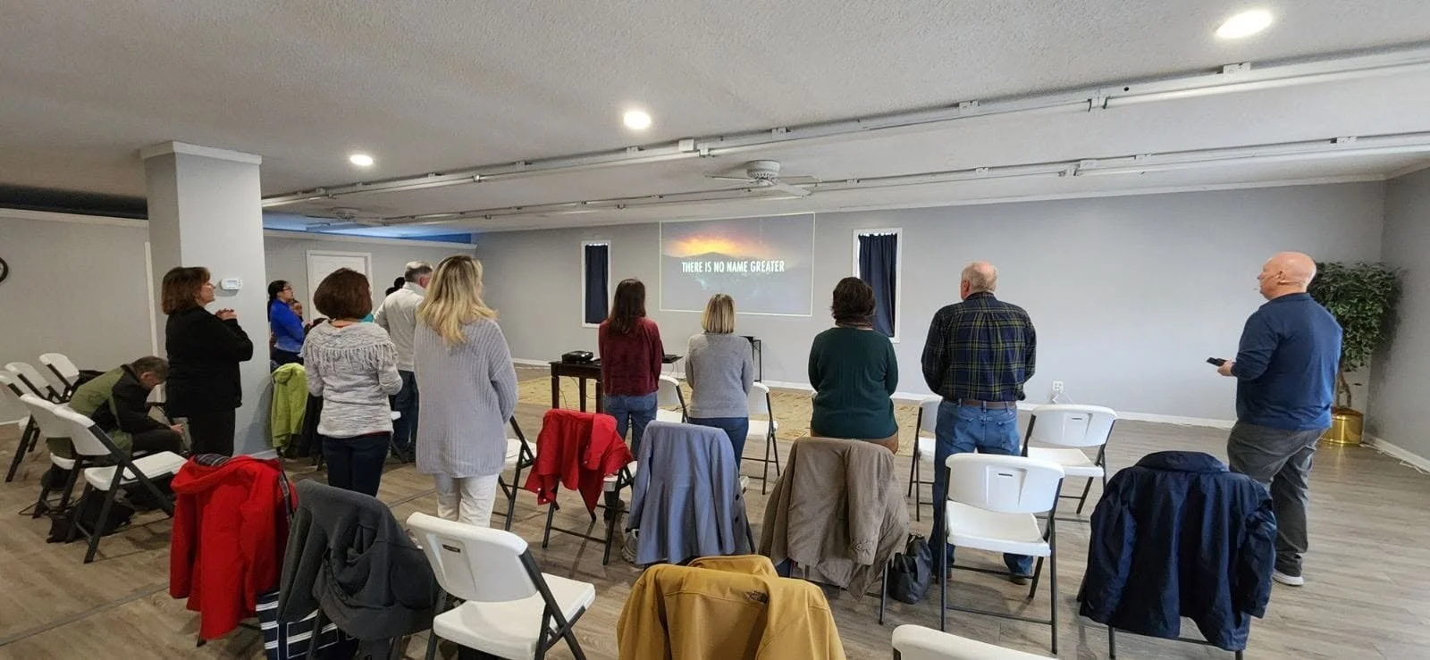 People standing and seated in a room watching a presentation with a screen that reads 'There is no name greater'.