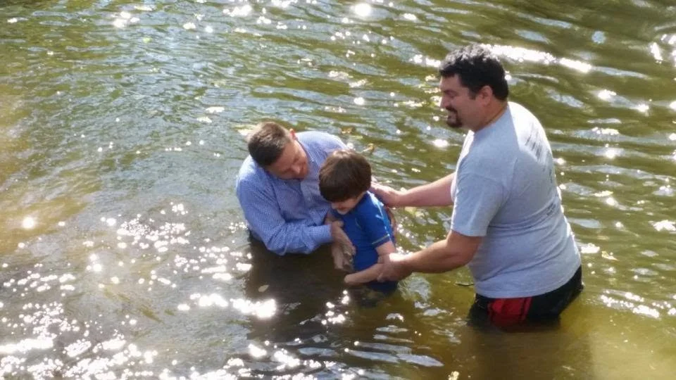 Three people, a man in a gray T-shirt, a boy in a blue shirt, and a man in a light blue shirt, are standing in a shallow river, with sunlight reflecting on the water. The man in the gray T-shirt is helping the boy in the blue shirt during a baptism or similar water ceremony.