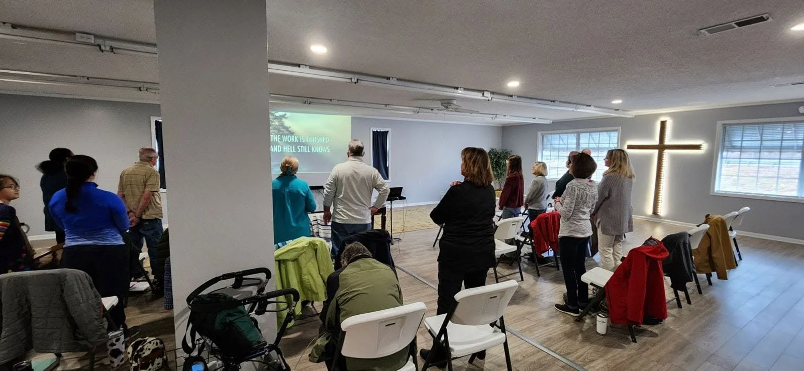 A group of people standing in a church or religious gathering space, facing a screen displaying a presentation. There is a large illuminated cross on the wall near the windows, and some chairs and strollers are visible in the room.