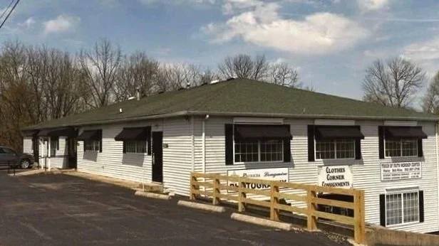 A single-story commercial building with a white exterior and black awnings over the windows, green roof, surrounded by a parking lot and a wooden fence, with trees in the background.
