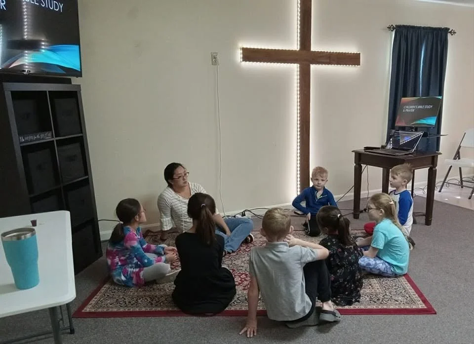 Children and a woman sitting cross-legged on a prayer rug in front of a large illuminated cross in a room, possibly a church or classroom, with a water cooler, a piano, a pull-down screen, and window with dark curtains.