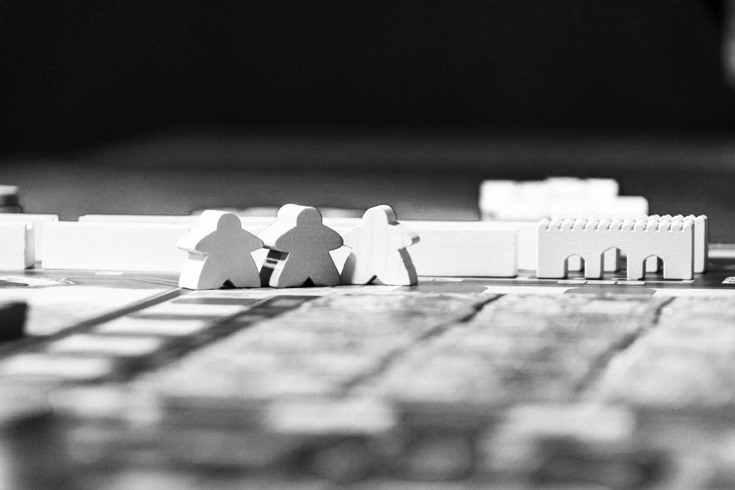 A black and white close-up of a section of a board game, showing three white game pieces shaped like human figures and some game pieces in the background.