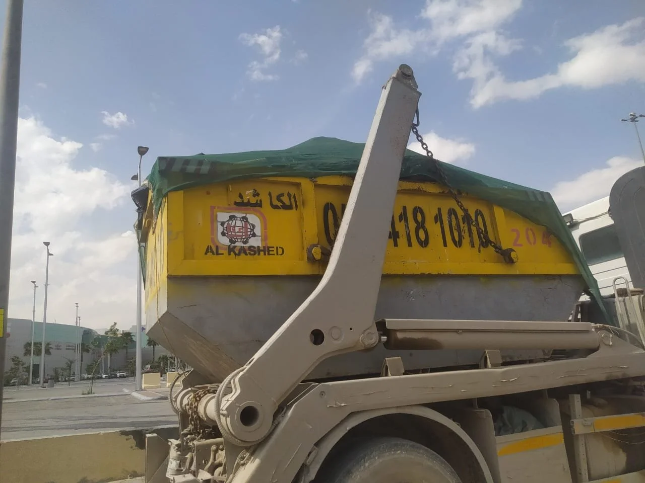 A construction dump truck with a yellow container, covered with a green tarp, parked in a parking lot with streetlights and a shopping mall in the background, under a partly cloudy sky.