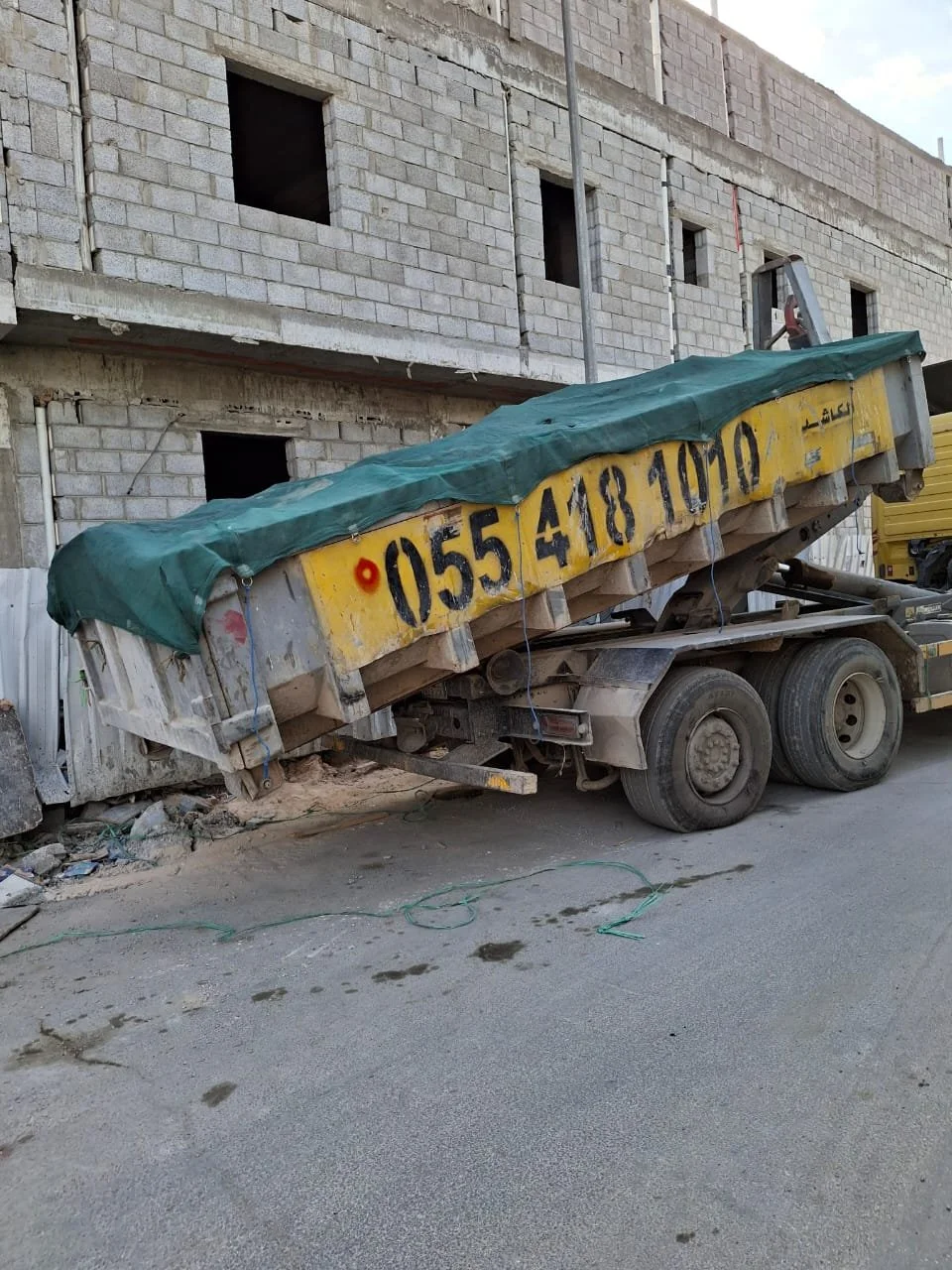 A construction dump truck parked on a street next to an unfinished brick building, with visible construction materials and debris on the ground.