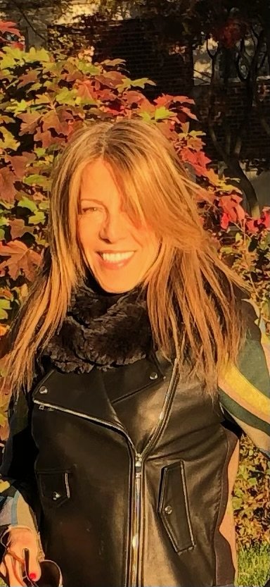 A smiling woman with long blonde hair in a black leather jacket, standing outdoors among autumn-colored leaves.