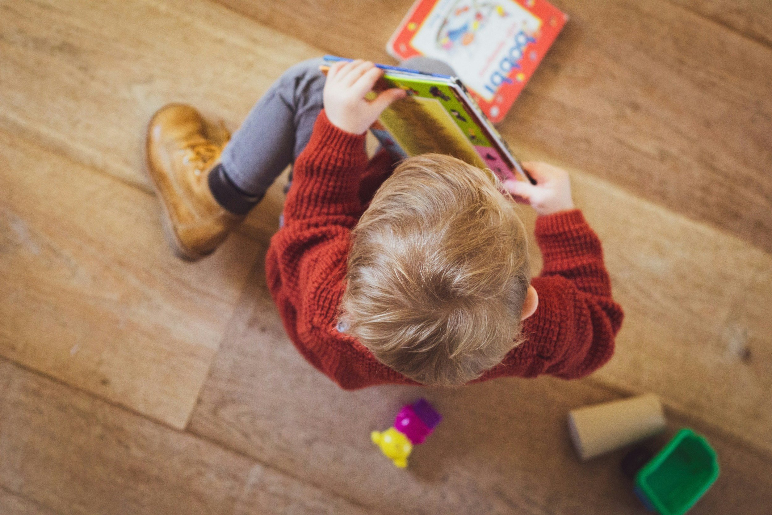 A child with light brown hair wearing a red sweater, gray pants, and tan boots, playing on a wooden floor with colorful toys and a board game nearby.