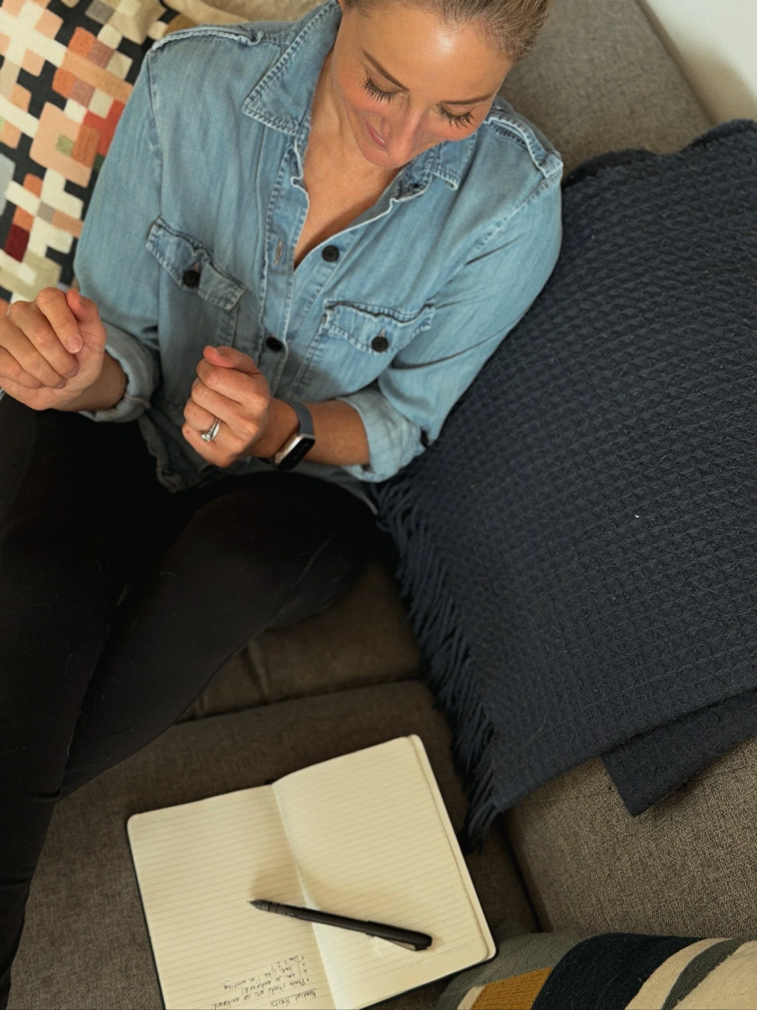 A woman sitting on a gray couch with a blue blanket, looking down with a notebook and a pen in front of her. There is a colorful pillow behind her and a person wearing striped clothing is partially visible on the right.