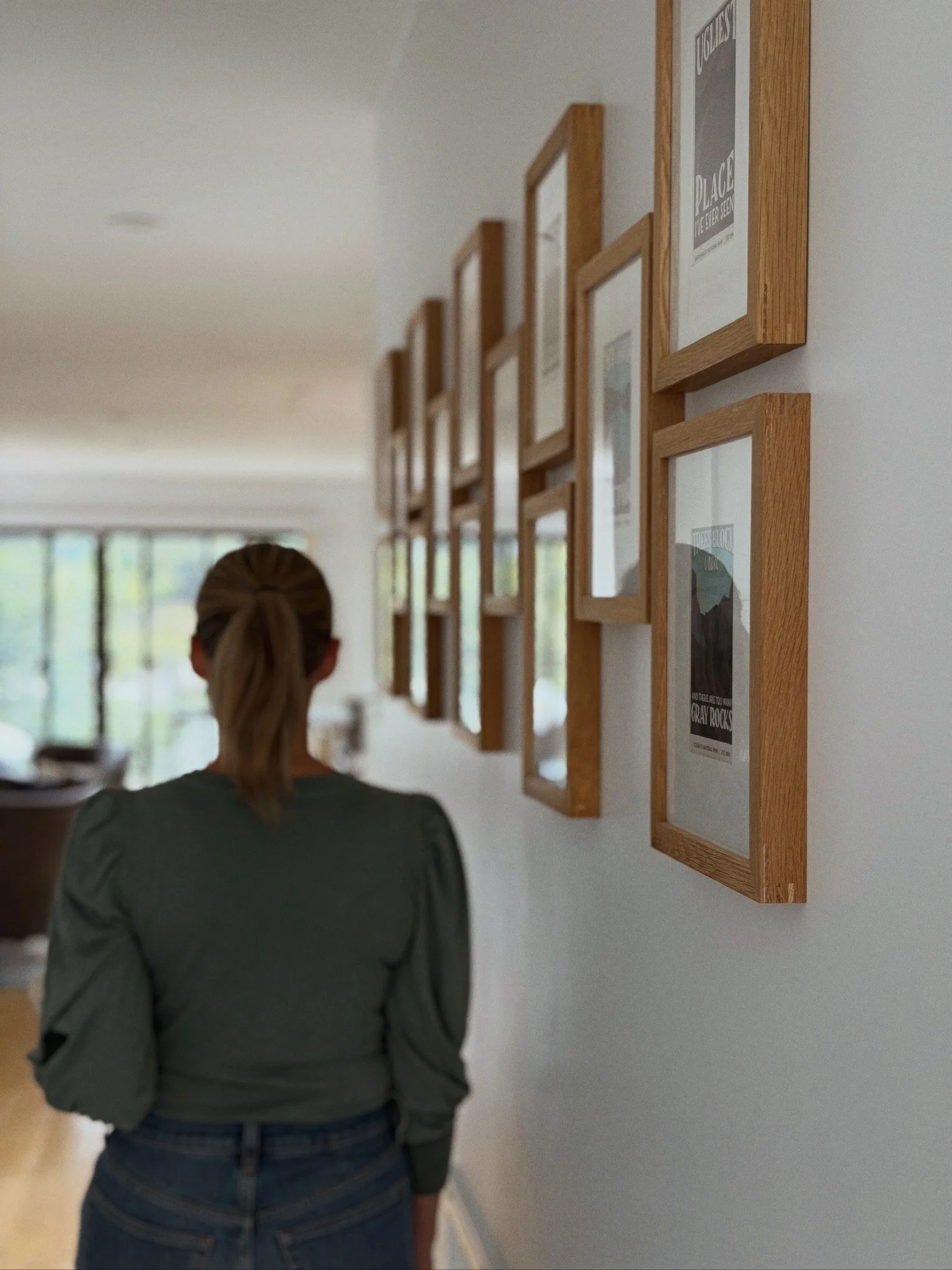 A woman with blonde hair tied back, wearing a gray long-sleeve shirt and blue jeans, stands with her back to the camera, observing framed posters on a white wall in a well-lit room with large windows.