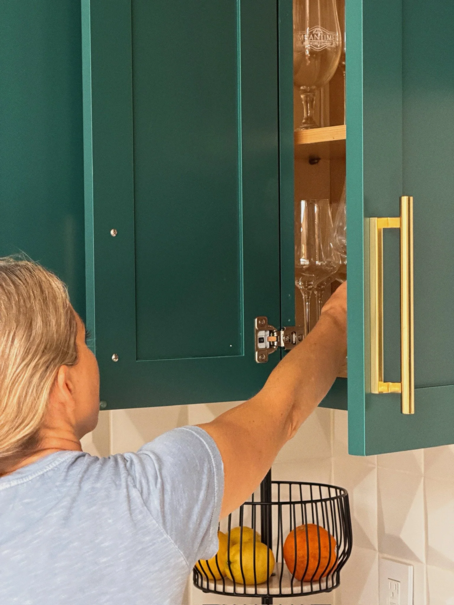 A woman with blonde hair, wearing a light blue shirt, is reaching into a teal cabinet in a kitchen. Inside the cabinet, a large glass trophy is visible. Below, on the kitchen counter, there is a black wire basket filled with yellow, orange, and brown decorative pumpkins.