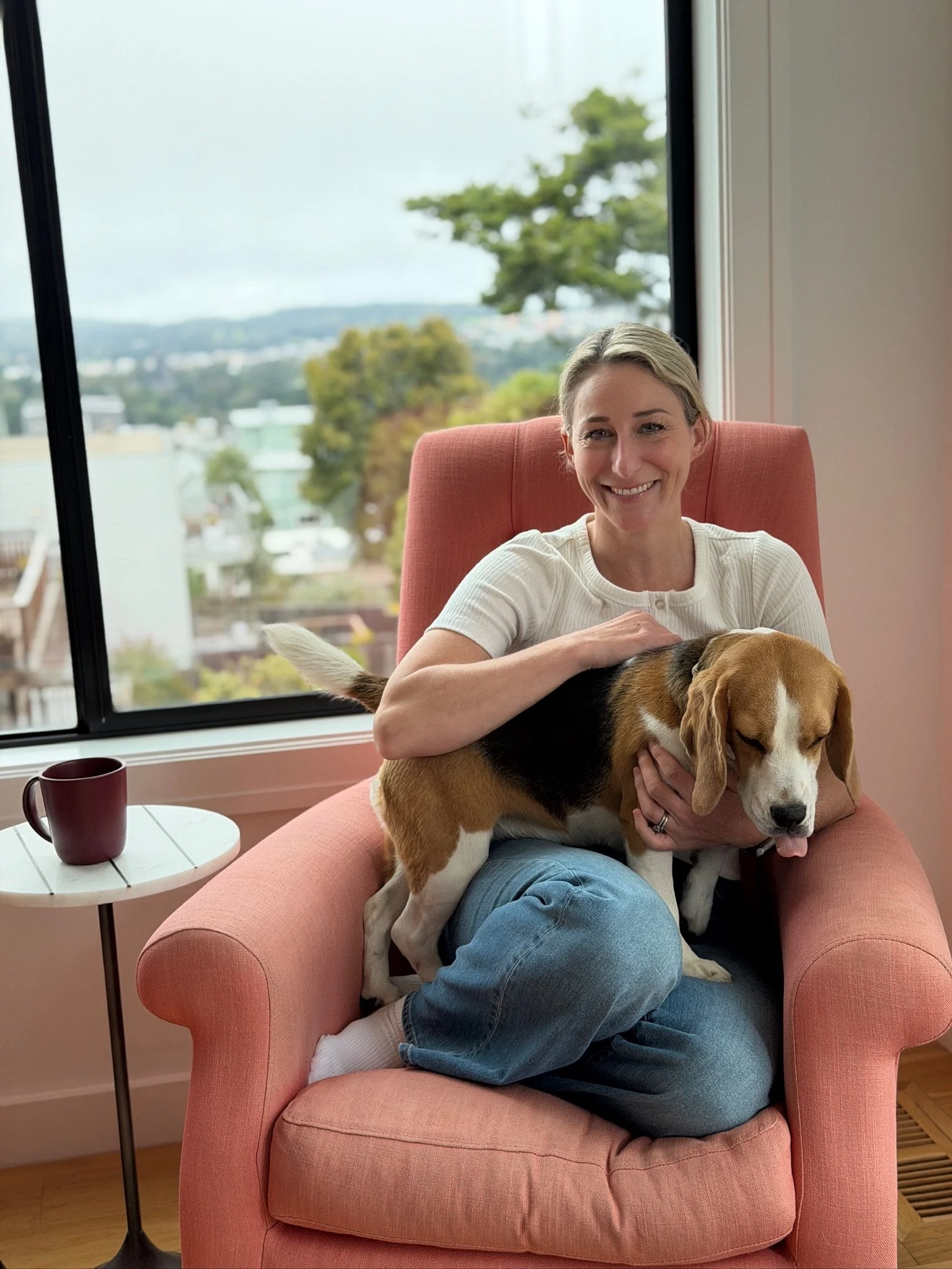 A woman with blonde hair smiling and sitting in a pink armchair, holding a beagle dog on her lap. The dog appears relaxed with its eyes closed. There is a window behind them showing a view of trees and city buildings.