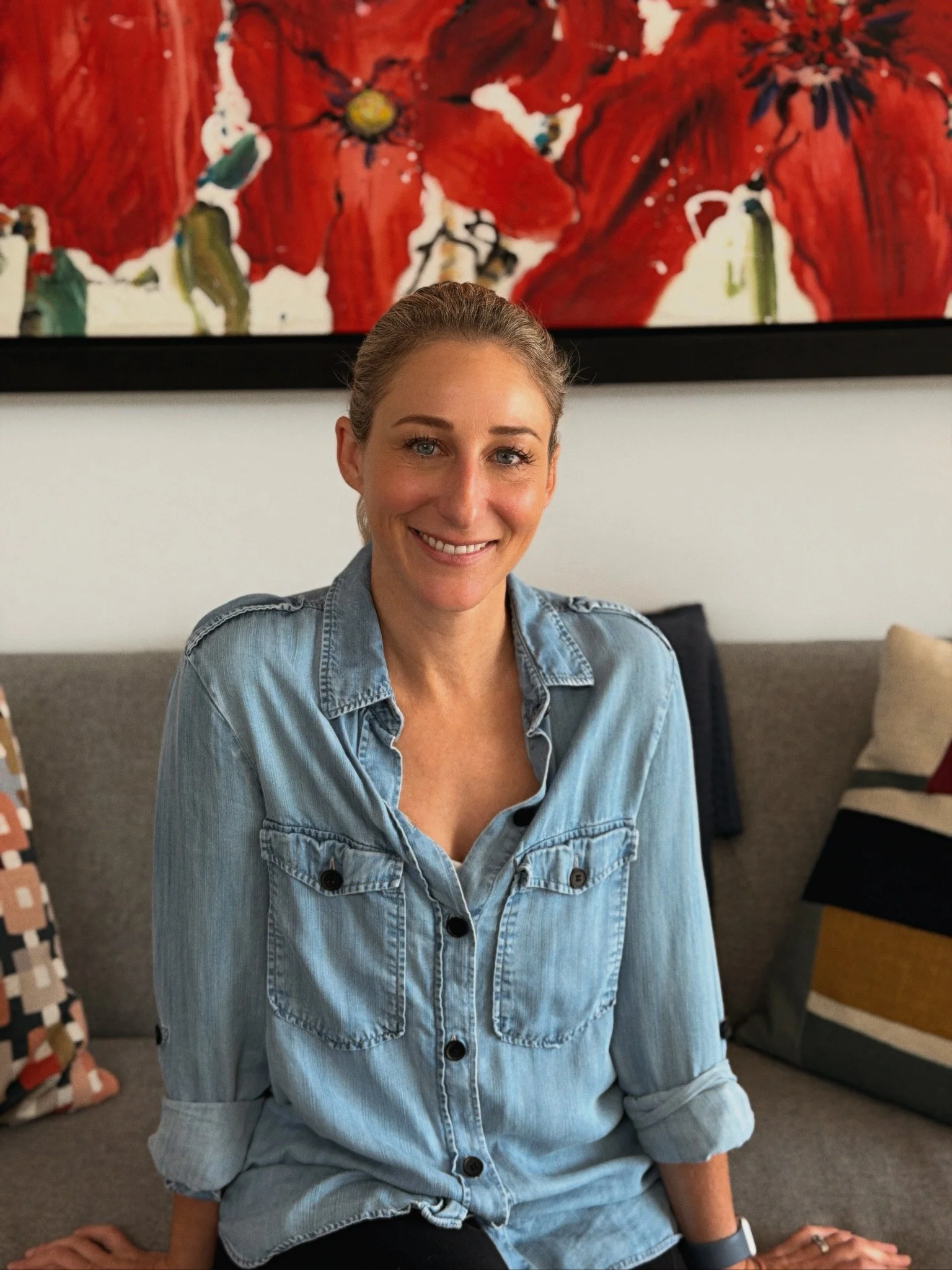 A woman with light brown hair pulled back, smiling and sitting on a sofa in a denim shirt, in front of a red flower painting.