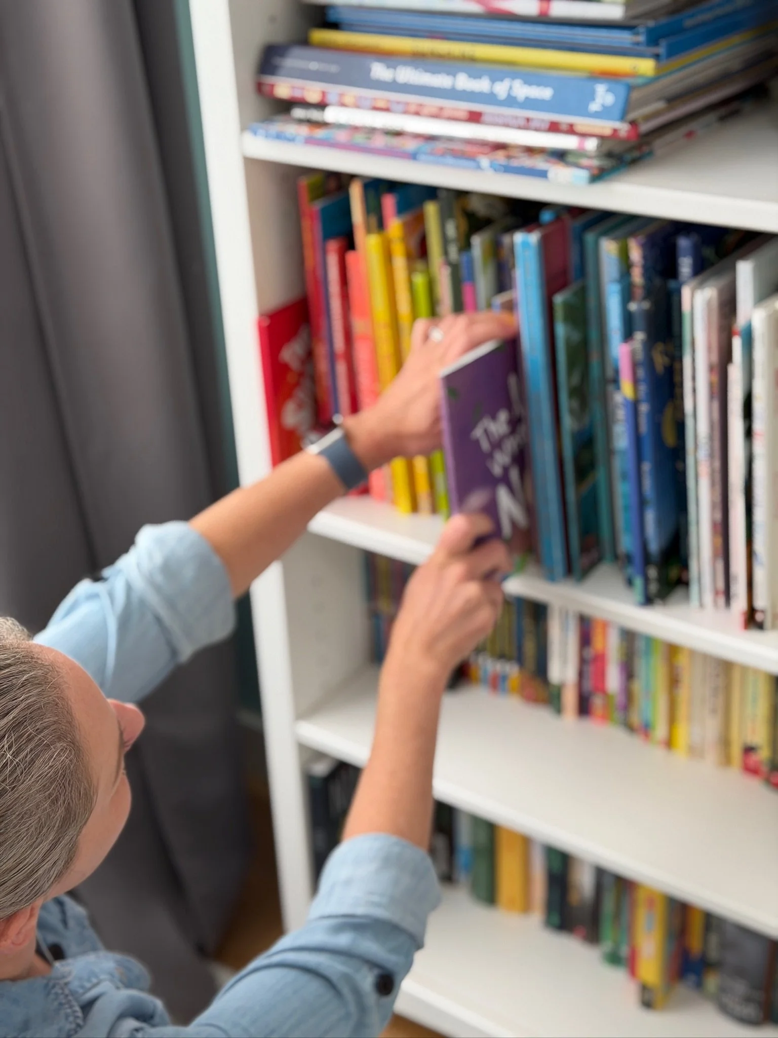 A person with gray hair, wearing a light blue denim shirt and a watch, reaching for a book on a white bookshelf filled with colorful books.