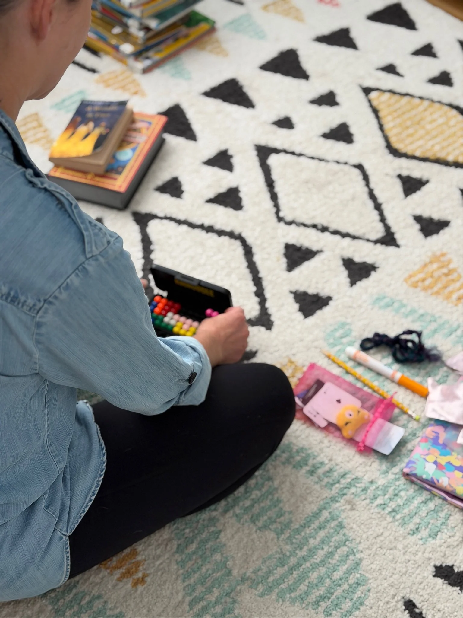 A person sitting on a patterned rug, playing a colorful game with small beads or counters, surrounded by various toys, books, and art supplies.