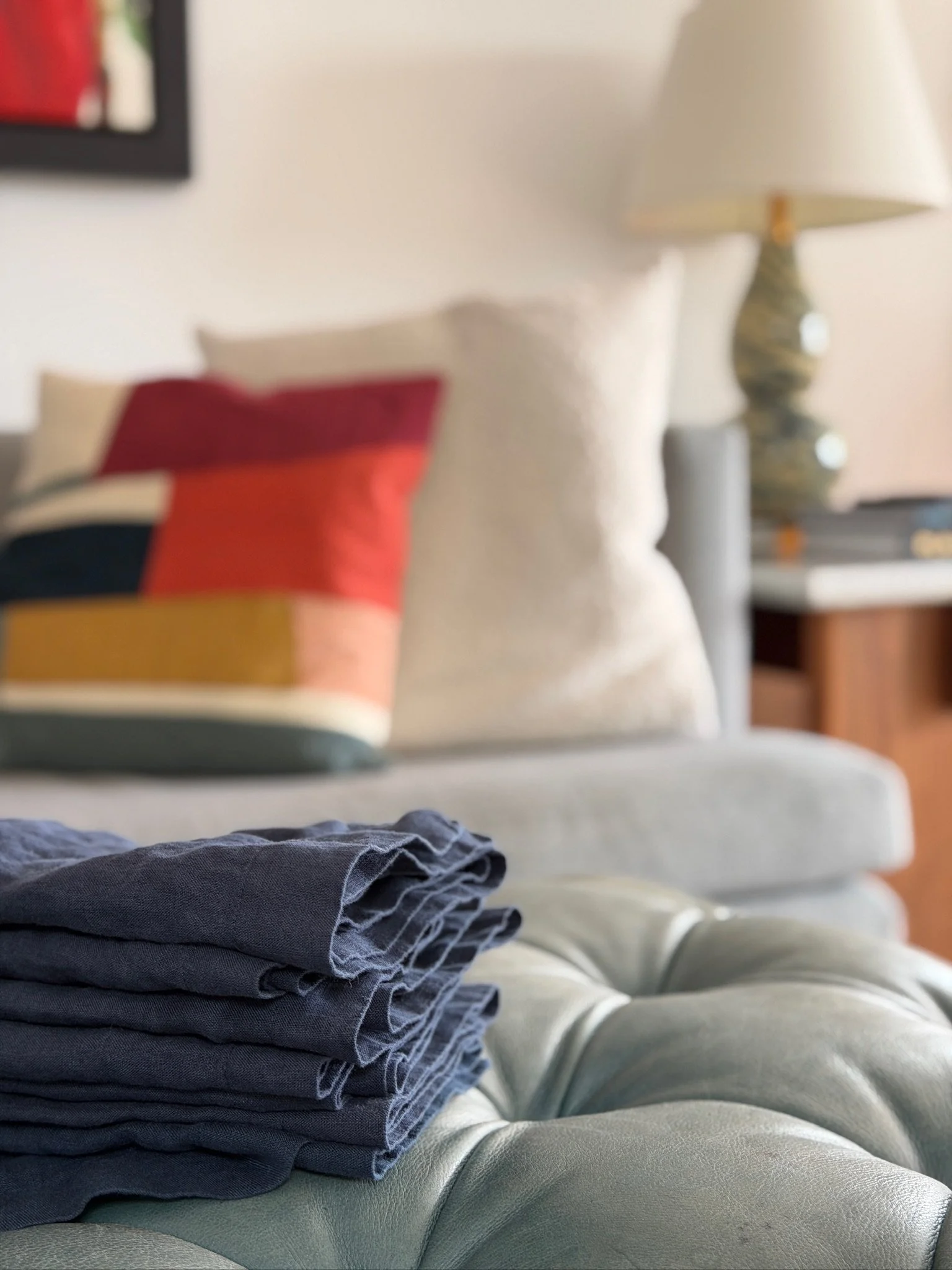 Stacked folded clothes on a light green sofa, with a background including a beige pillow, a table with a lamp, and a blurred picture on the wall.