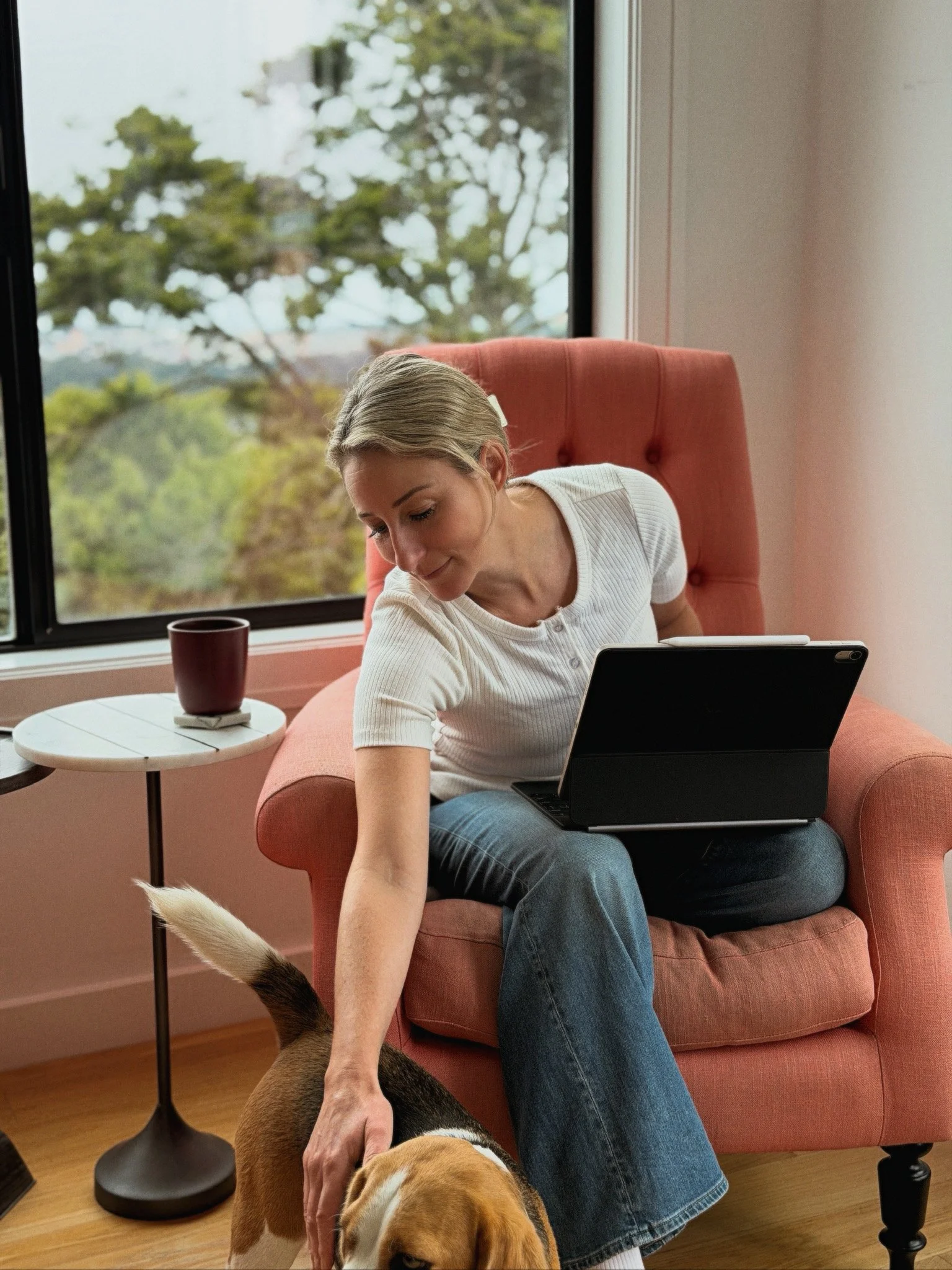 Woman sitting in an orange armchair, petting two dogs. A window with greenery outside is behind her, and a small round side table with a dark red mug is beside her.