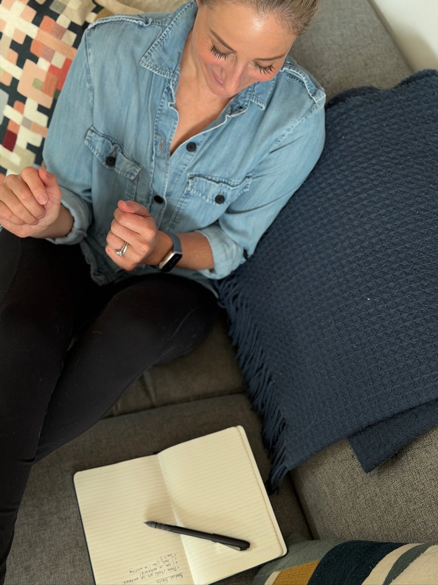 A person sitting on a sofa wearing a denim shirt, with an open notebook and a pen on the coffee table in front of them.